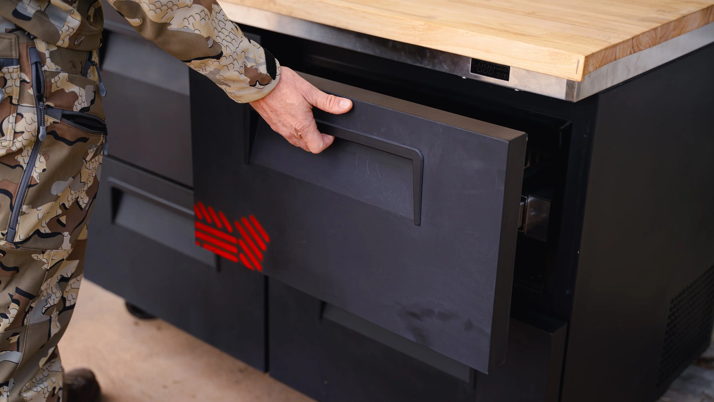 A person in camouflage clothing opening a black safe or drawer under a wooden countertop.