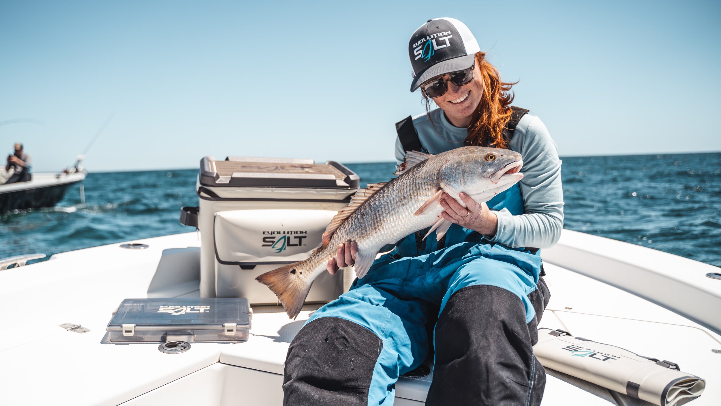 Woman on a boat holding a large fish, wearing sunglasses and a baseball cap, with other boaters fishing in the background.