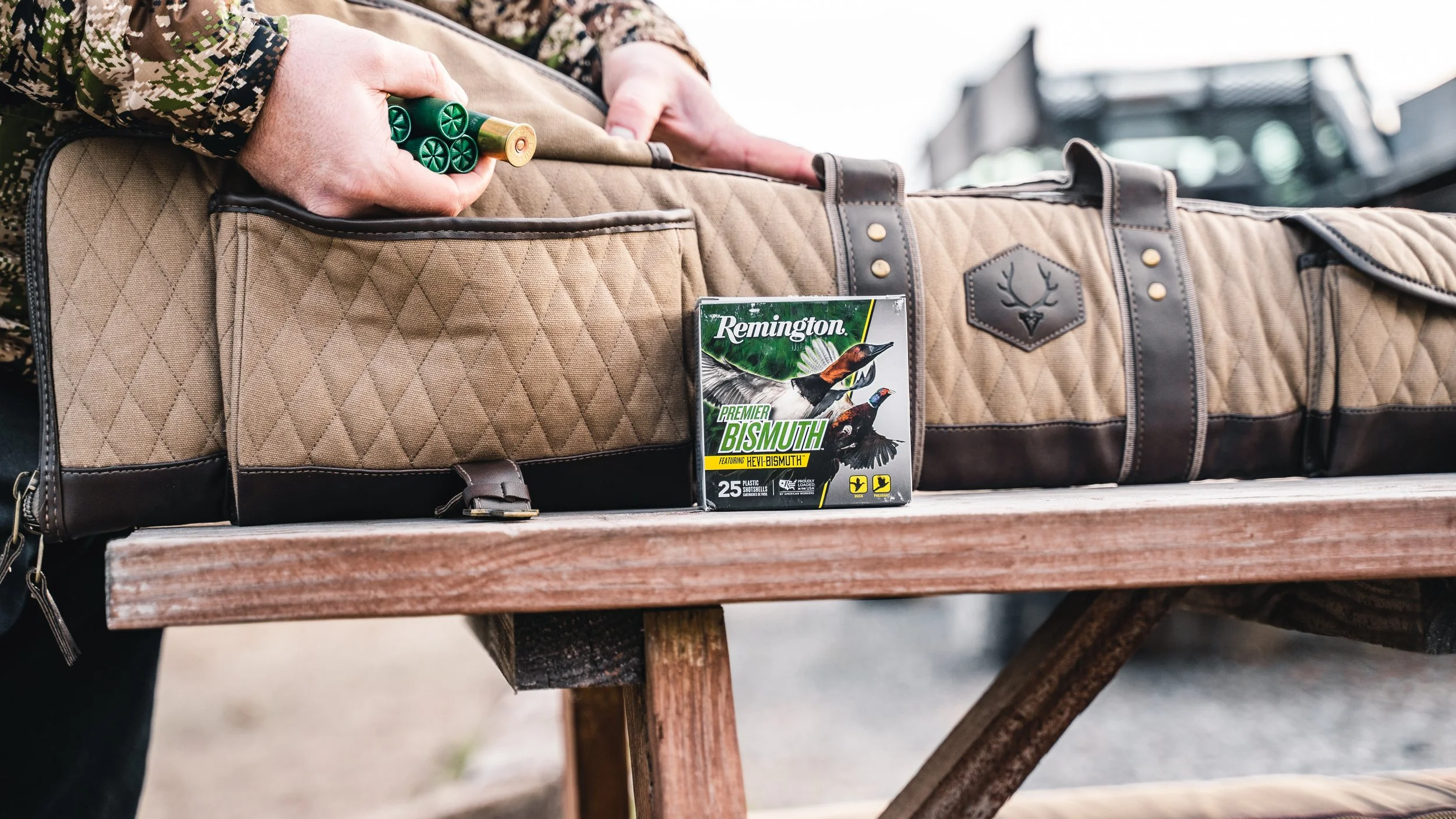 A person in camouflage clothing is placing a shotgun shell into a gun case. There is a box of Remington Premier Bismuth shotgun shells on the table, with pictures of birds on the front.