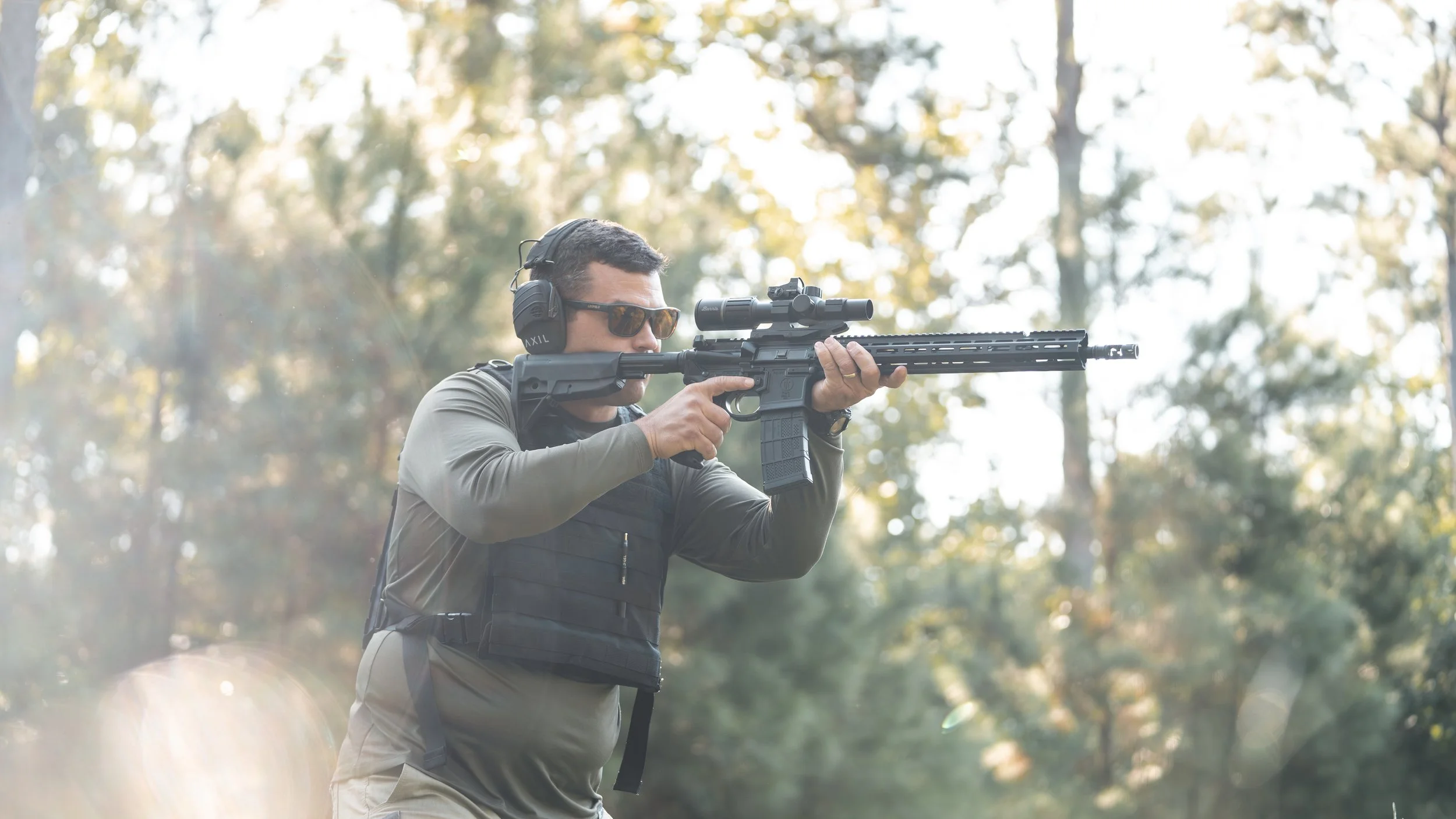 A man aiming a rifle with scope outdoors, wearing headphones, sunglasses, and tactical gear.