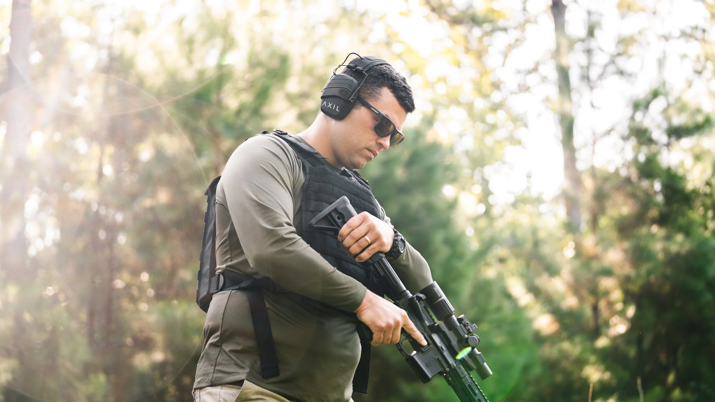 A man wearing sunglasses and ear protection inspects a rifle outdoors in a forested area during daylight.
