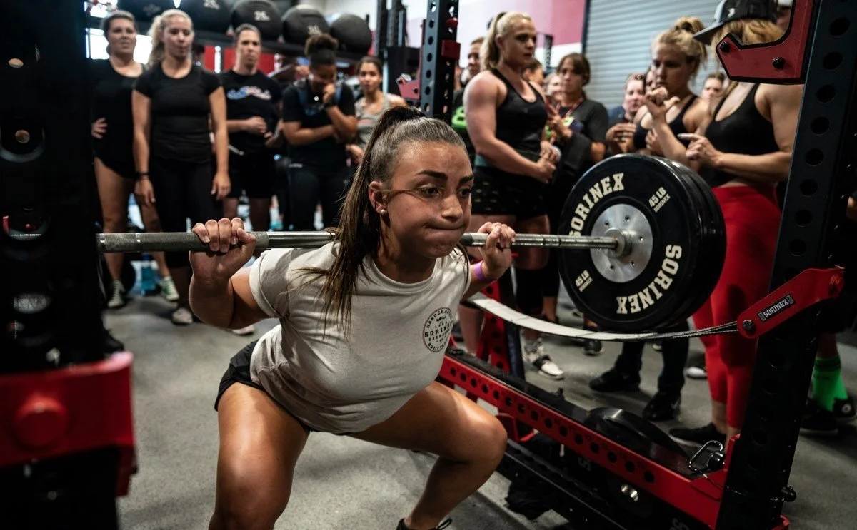 Woman performing a squat with a barbell in a gym, surrounded by spectators.