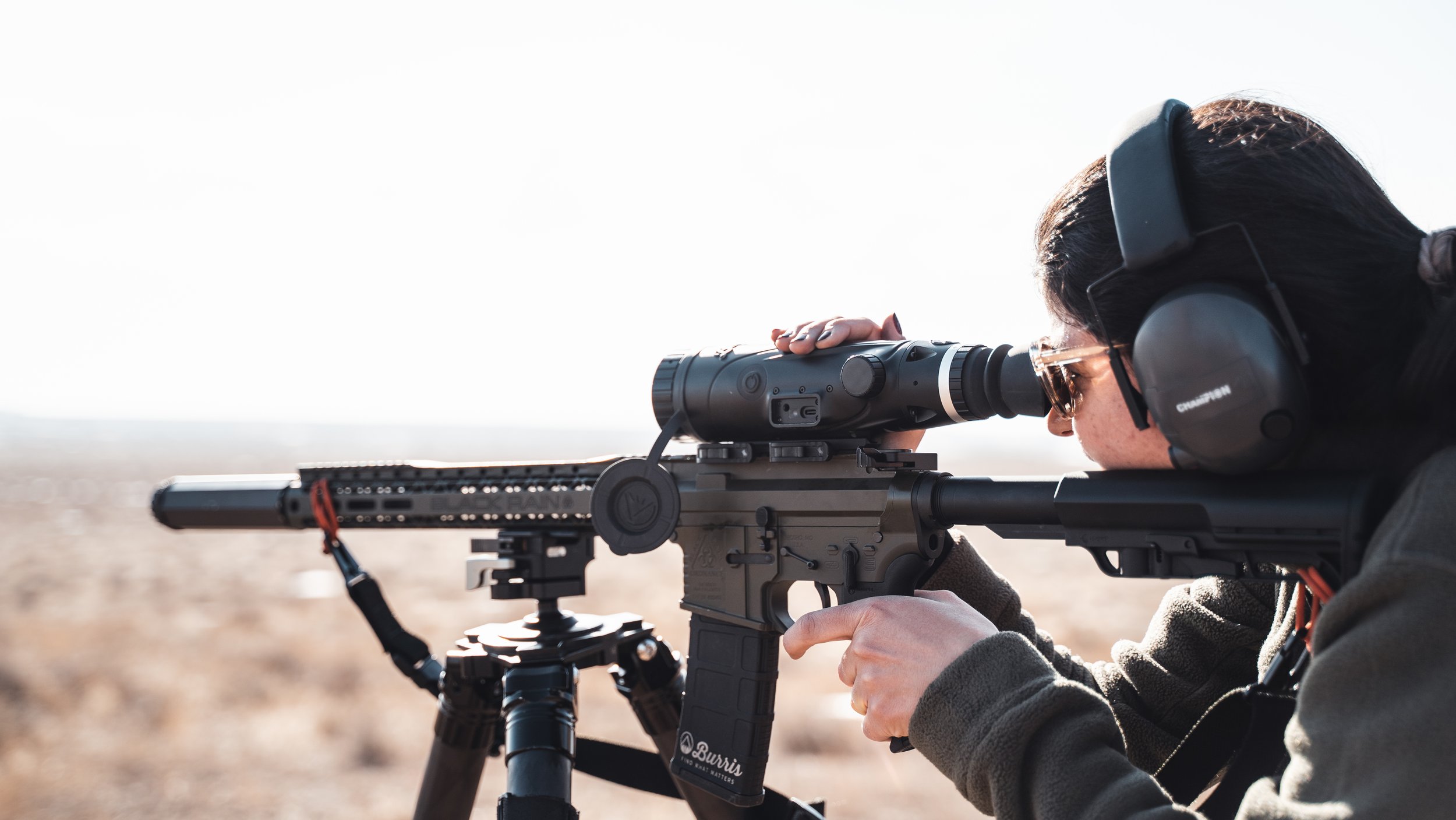 A woman aiming a sniper rifle with a scope in an outdoor desert landscape, wearing protective hearing earmuffs and sunglasses.