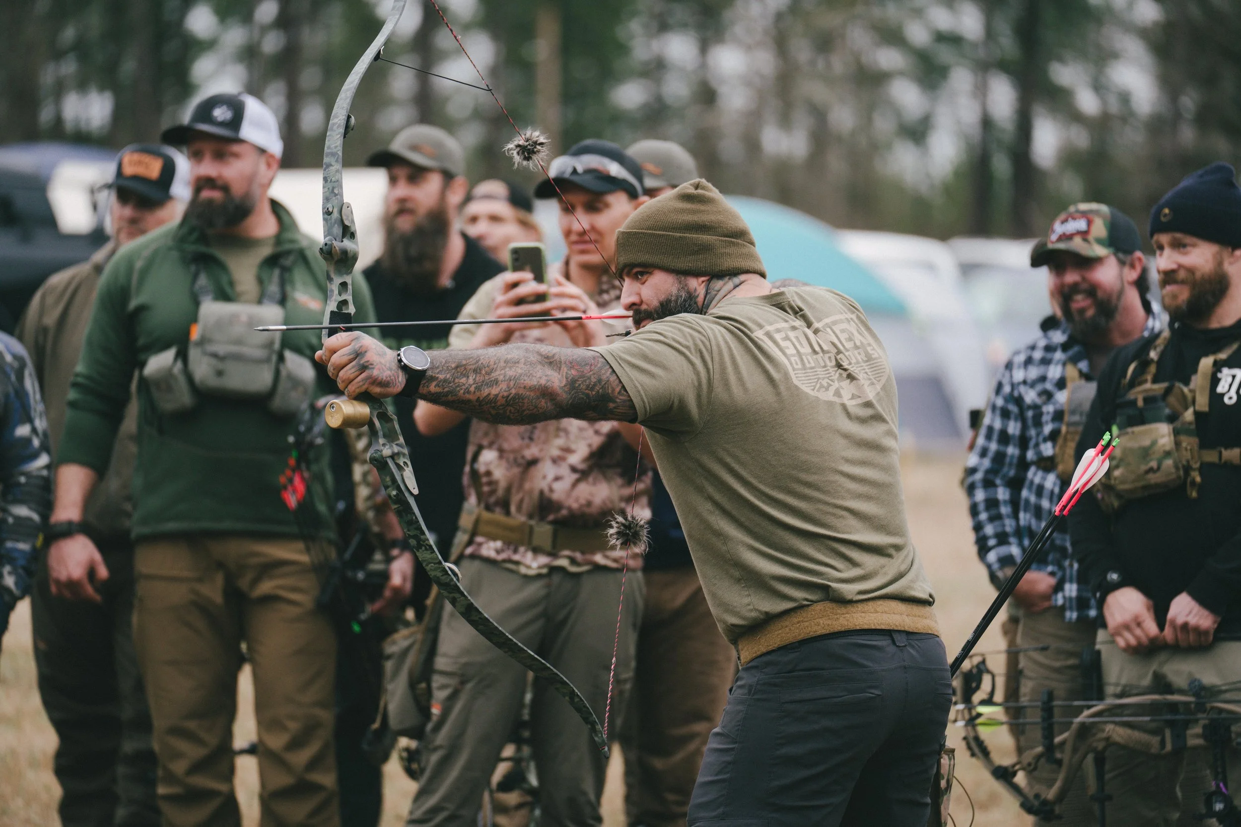 A man aiming a compound bow during an archery event with a group of people watching outdoors in a wooded area.