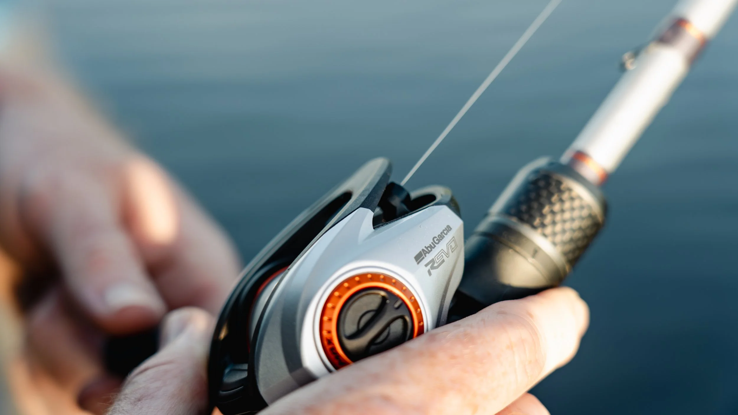 Close-up of a fishing reel and rod held by a person's hand, with water in the background.