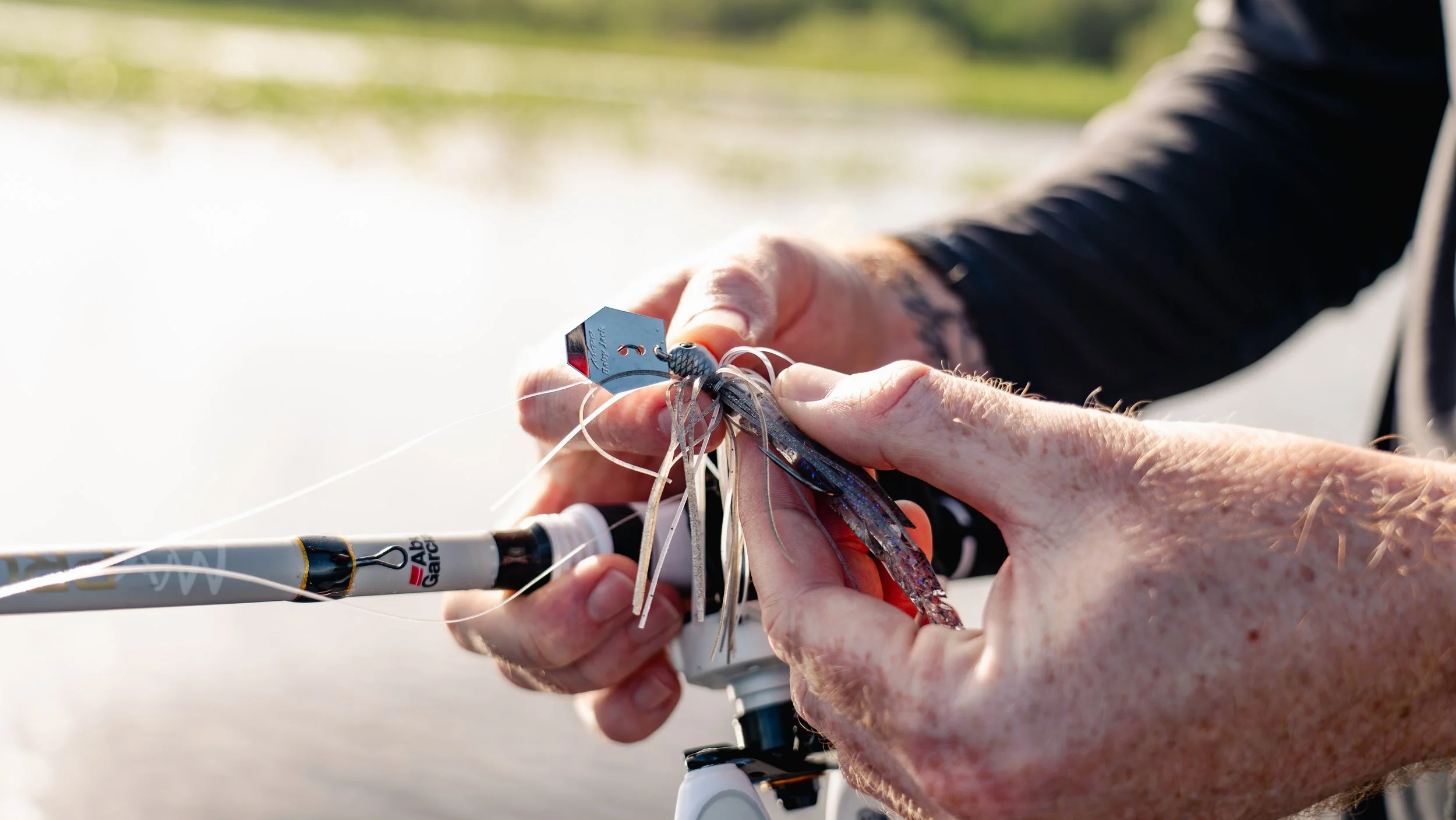 Close-up of a person's hands attaching a fishing lure to a fishing line near a body of water with a blurred background of green trees.