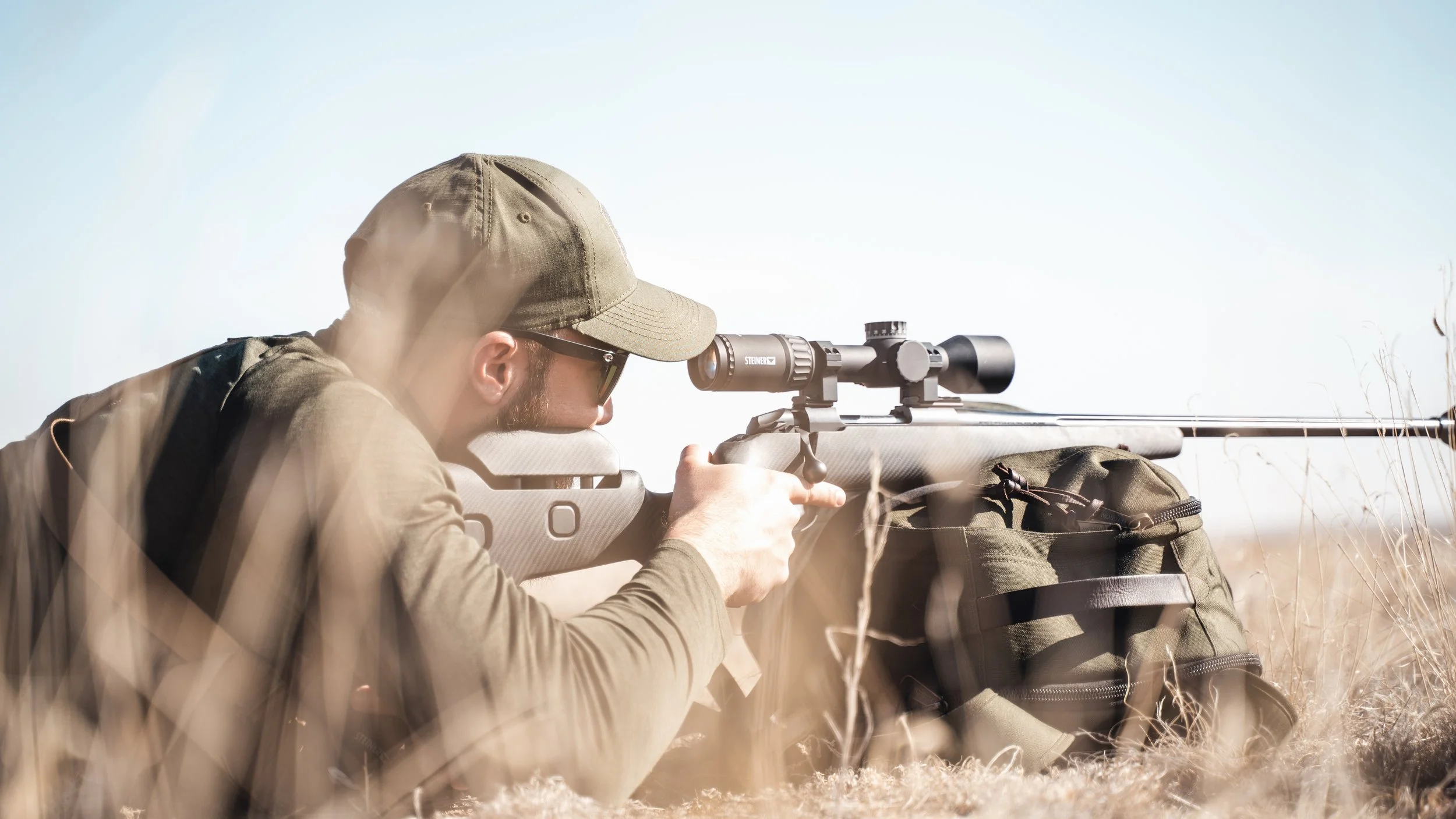 A man in a tan cap and sunglasses aiming a sniper rifle with a scope, laying prone on the ground in dry grass, with a backpack nearby, under a clear sky.