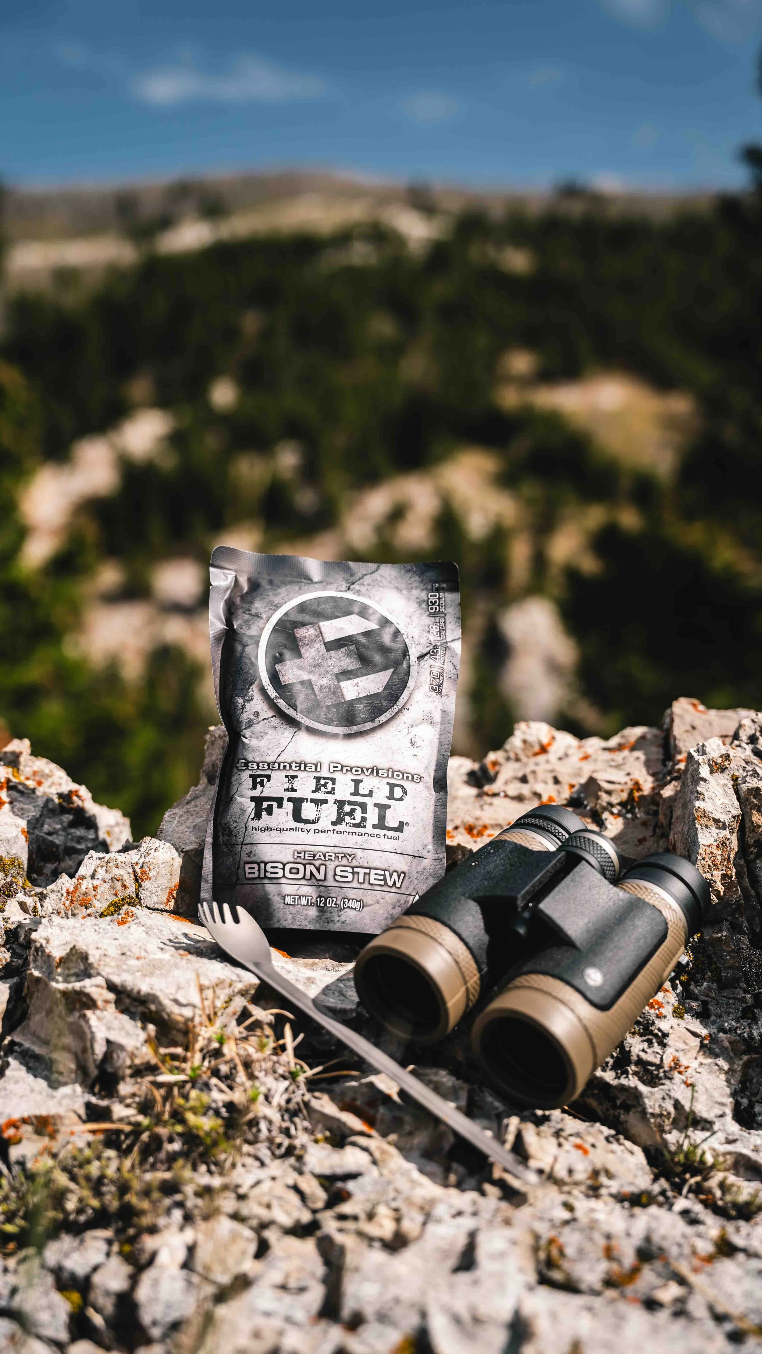 A packet of fuel, binoculars, and a small fork are placed on rocky terrain with a blurred mountainous landscape in the background.