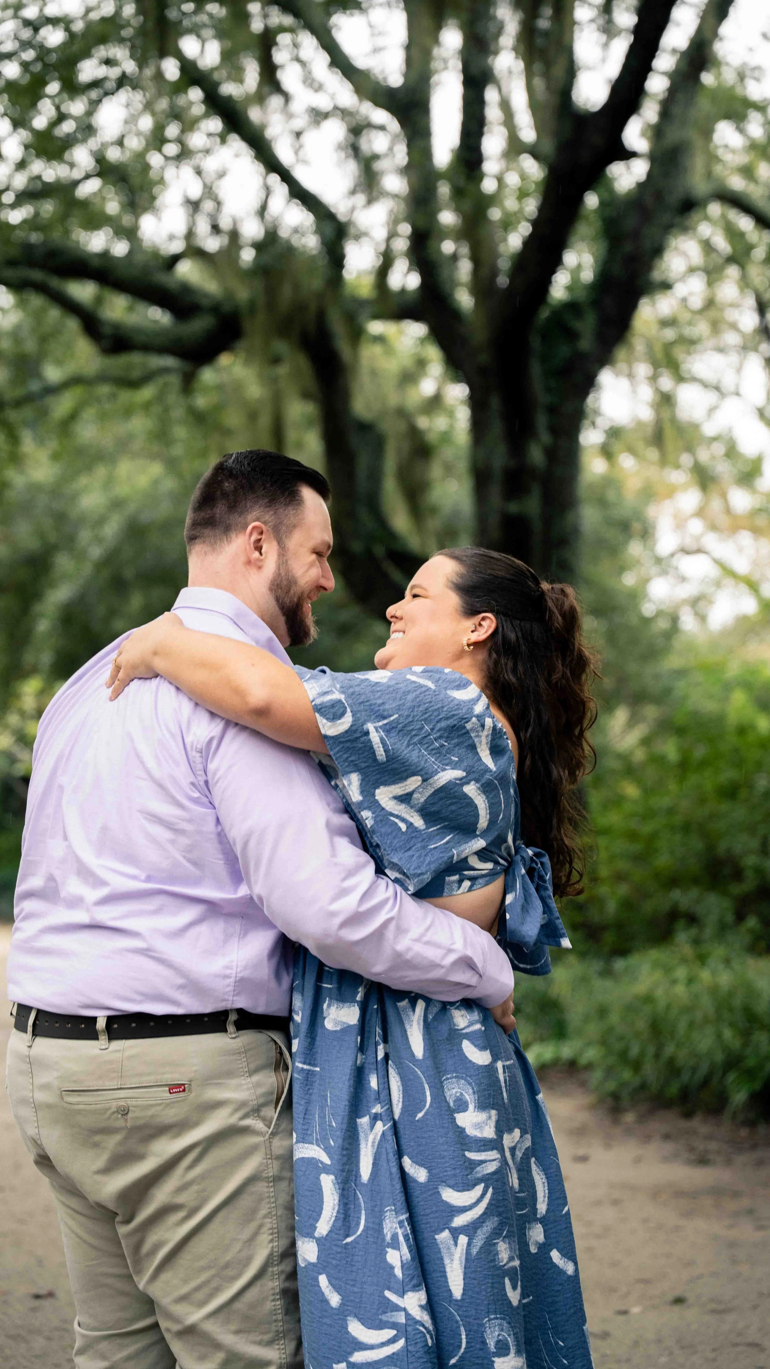 A couple embraces and smiles at each other in a park with lush green trees in the background.