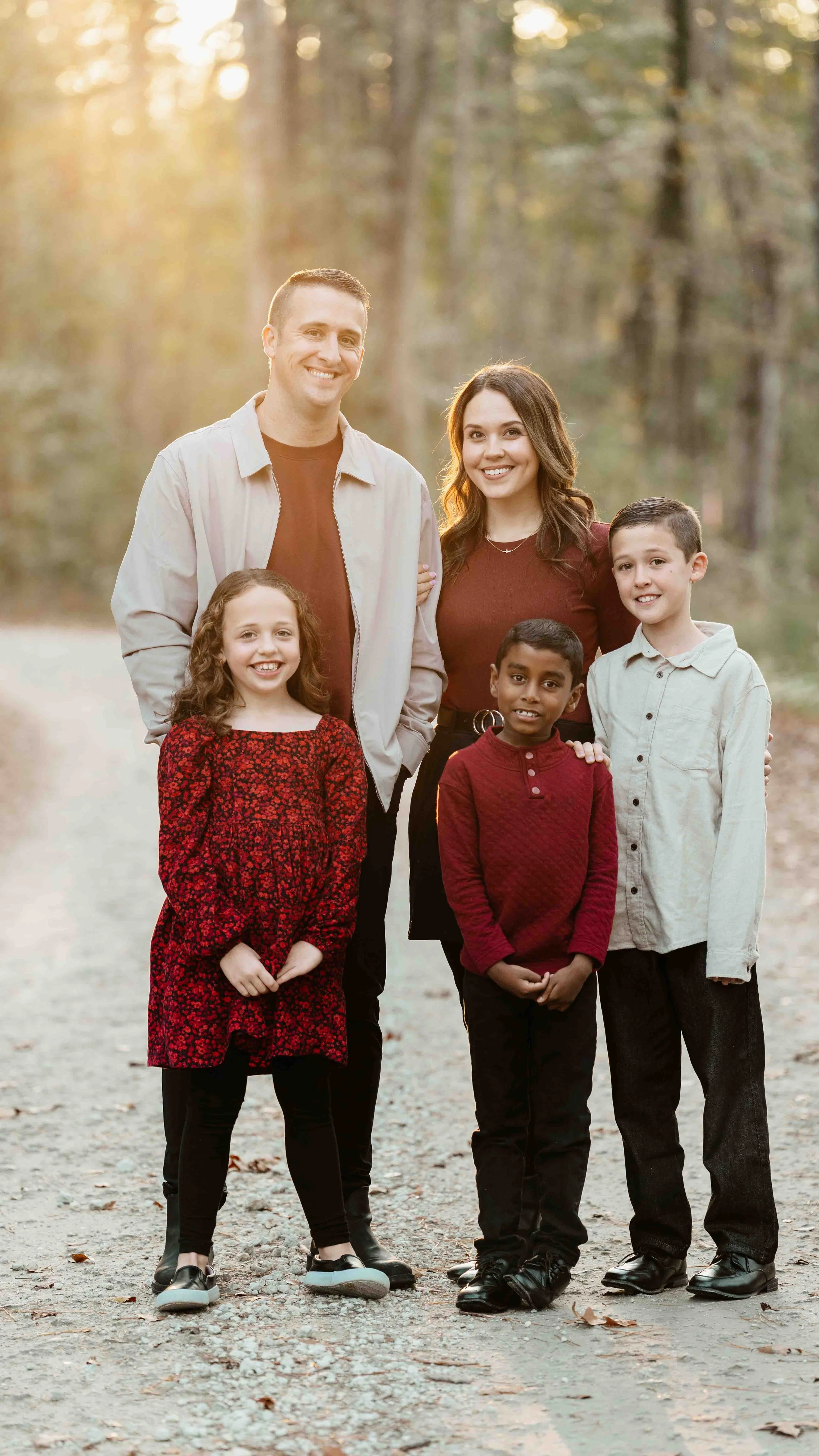 A family of six standing outdoors on a dirt path in a forested area during sunset, smiling at the camera.