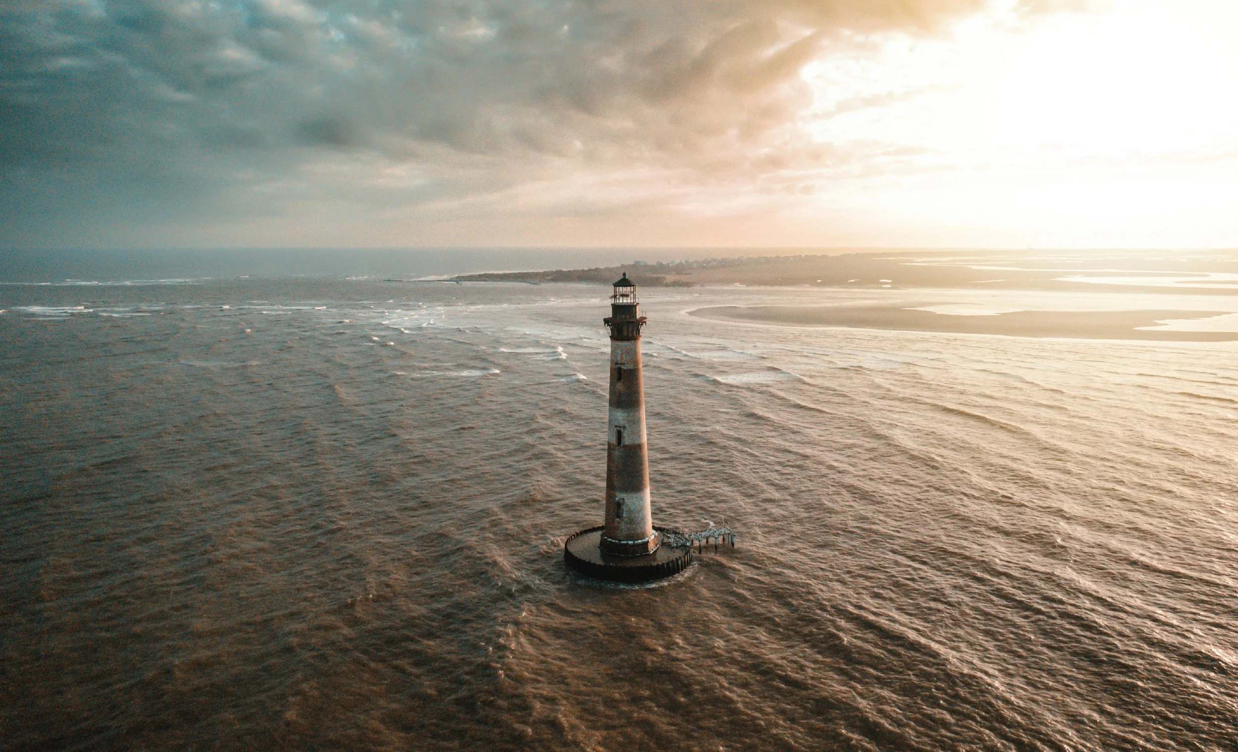 An aerial view of a lighthouse standing alone in the ocean near the coast during sunset or sunrise, with a cloudy sky and distant shoreline.