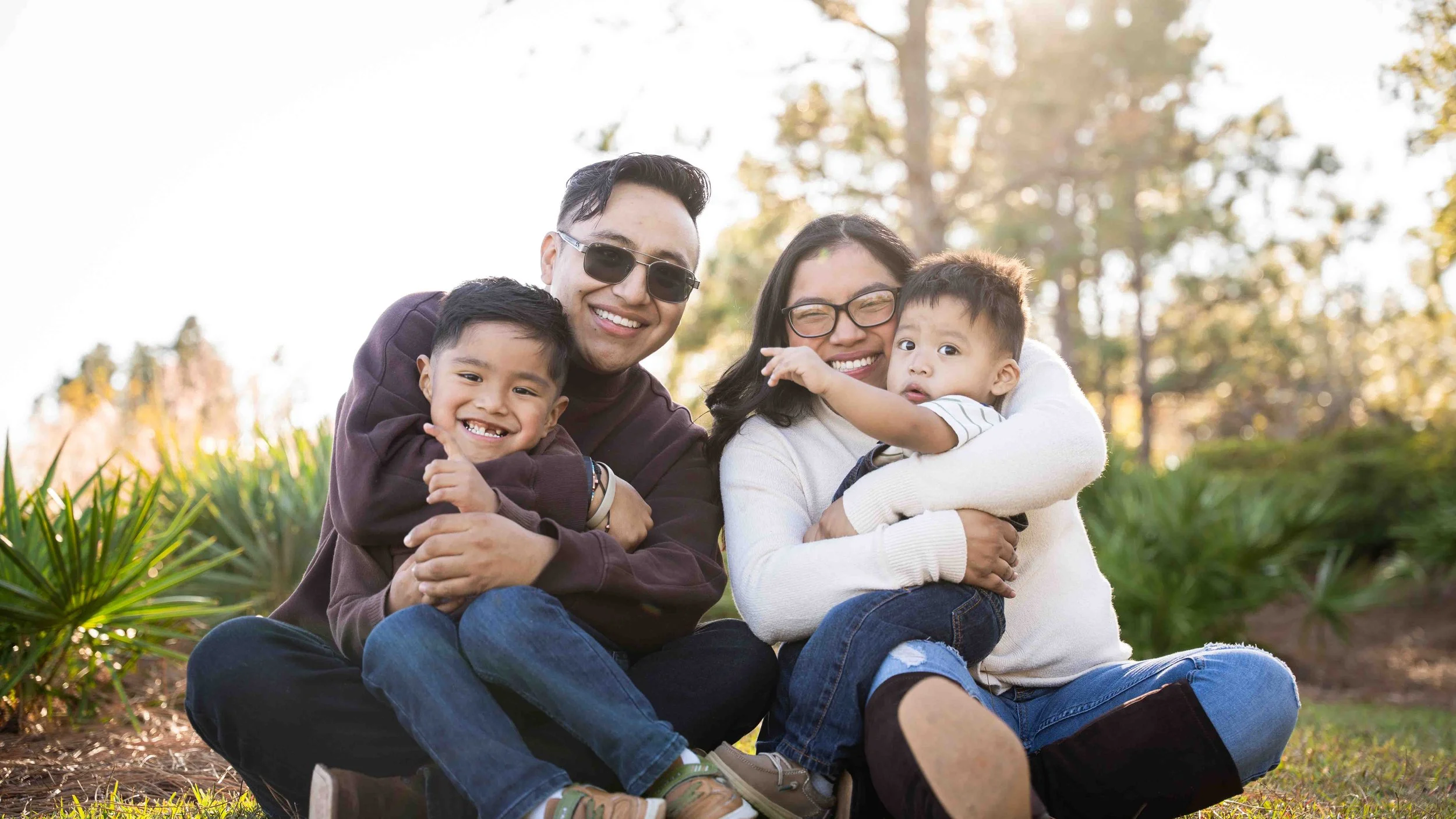 Four people, two adults and two children, sitting on grass outdoors, smiling and hugging each other, with trees and sunlight in the background.