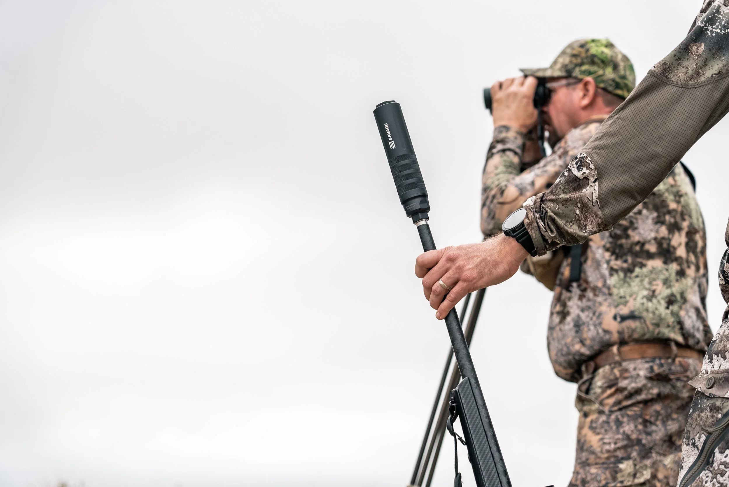 A person in camouflage clothing holding binoculars with another person operating some kind of surveying equipment with a microphone.