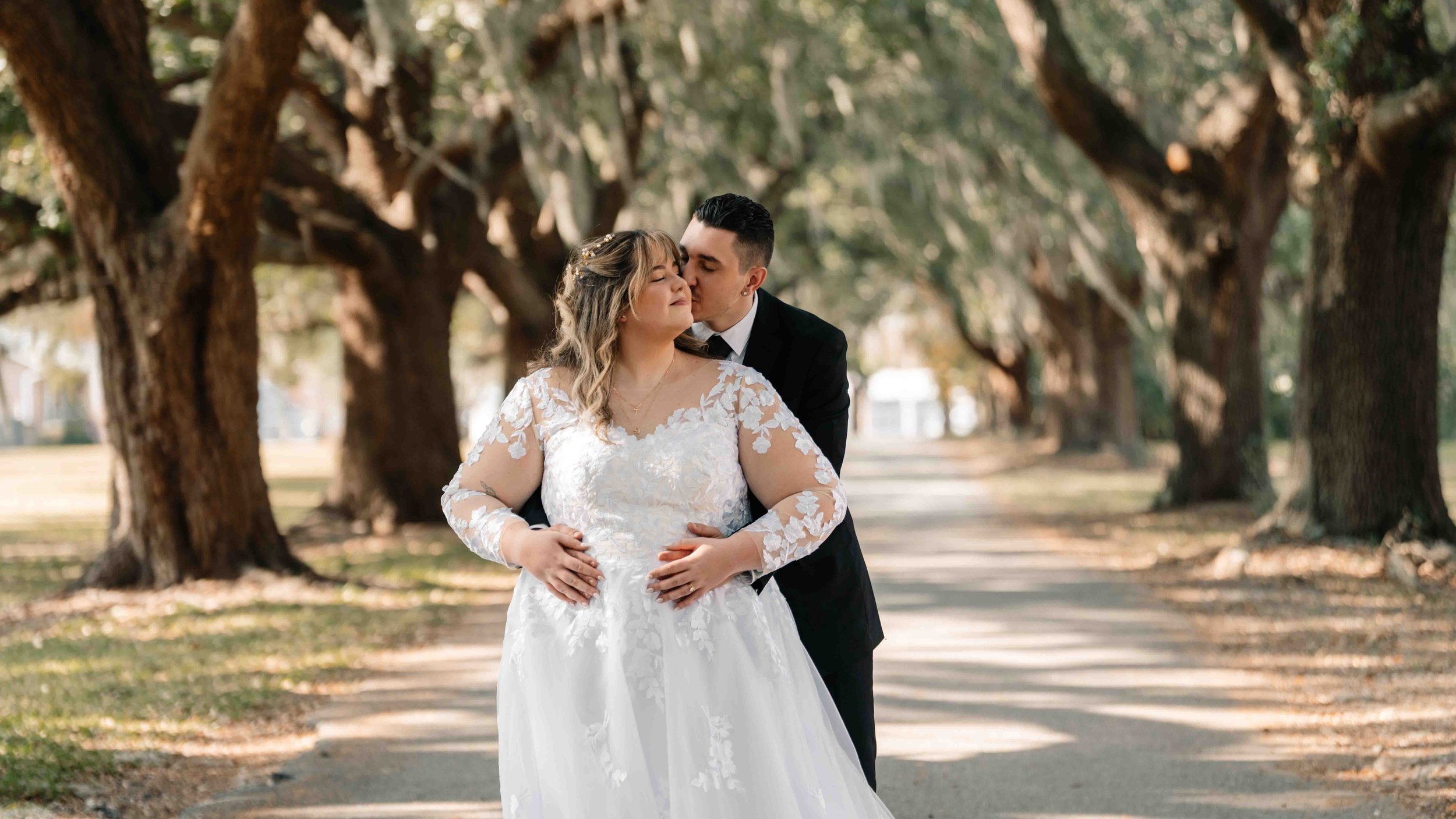 A bride in a white wedding dress with lace sleeves and a groom in a black suit share a romantic moment on a tree-lined path, with the groom embracing the bride from behind as they lean in close.