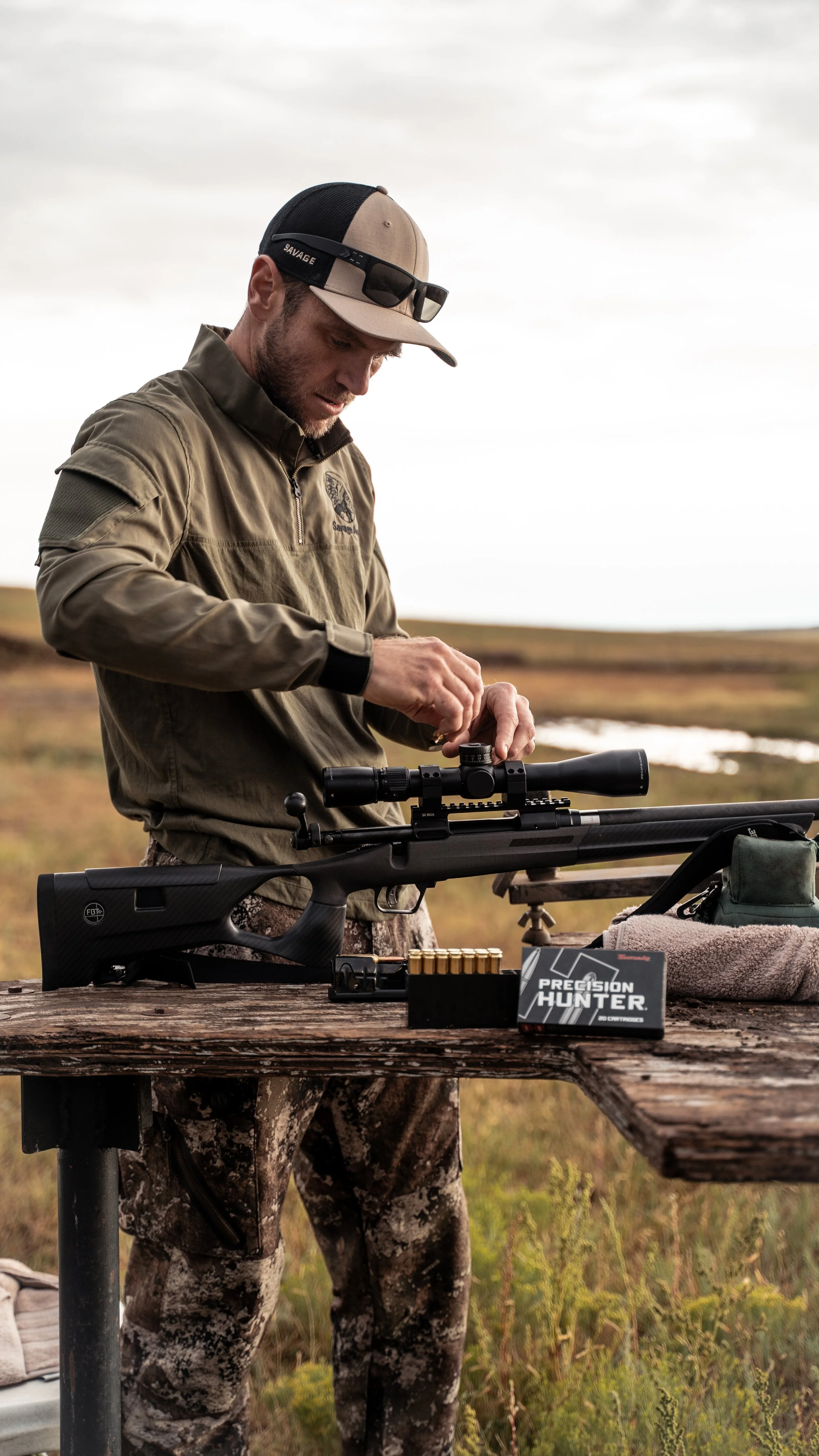 A man adjusting scope on a black rifle on a wooden table outdoors in a field.