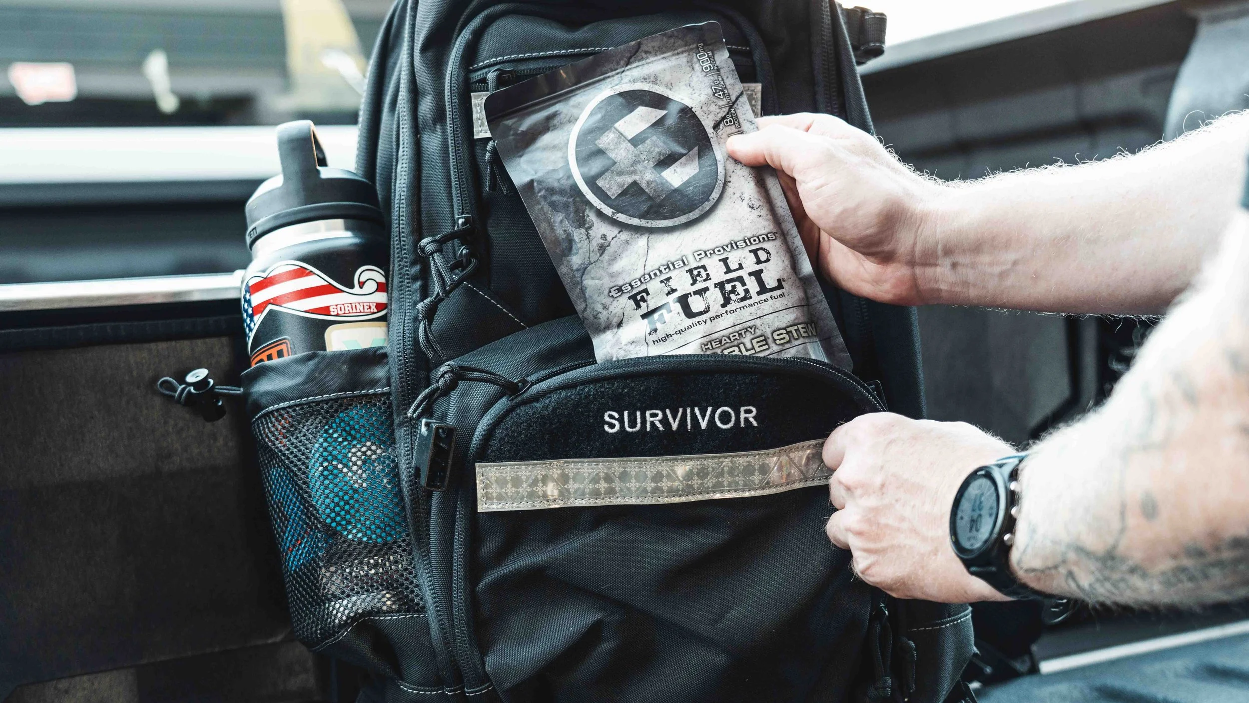 Person placing a packet of fuel labeled 'Hearty Double Stew' into a backpack with an 'SURVIVOR' patch, inside a vehicle.