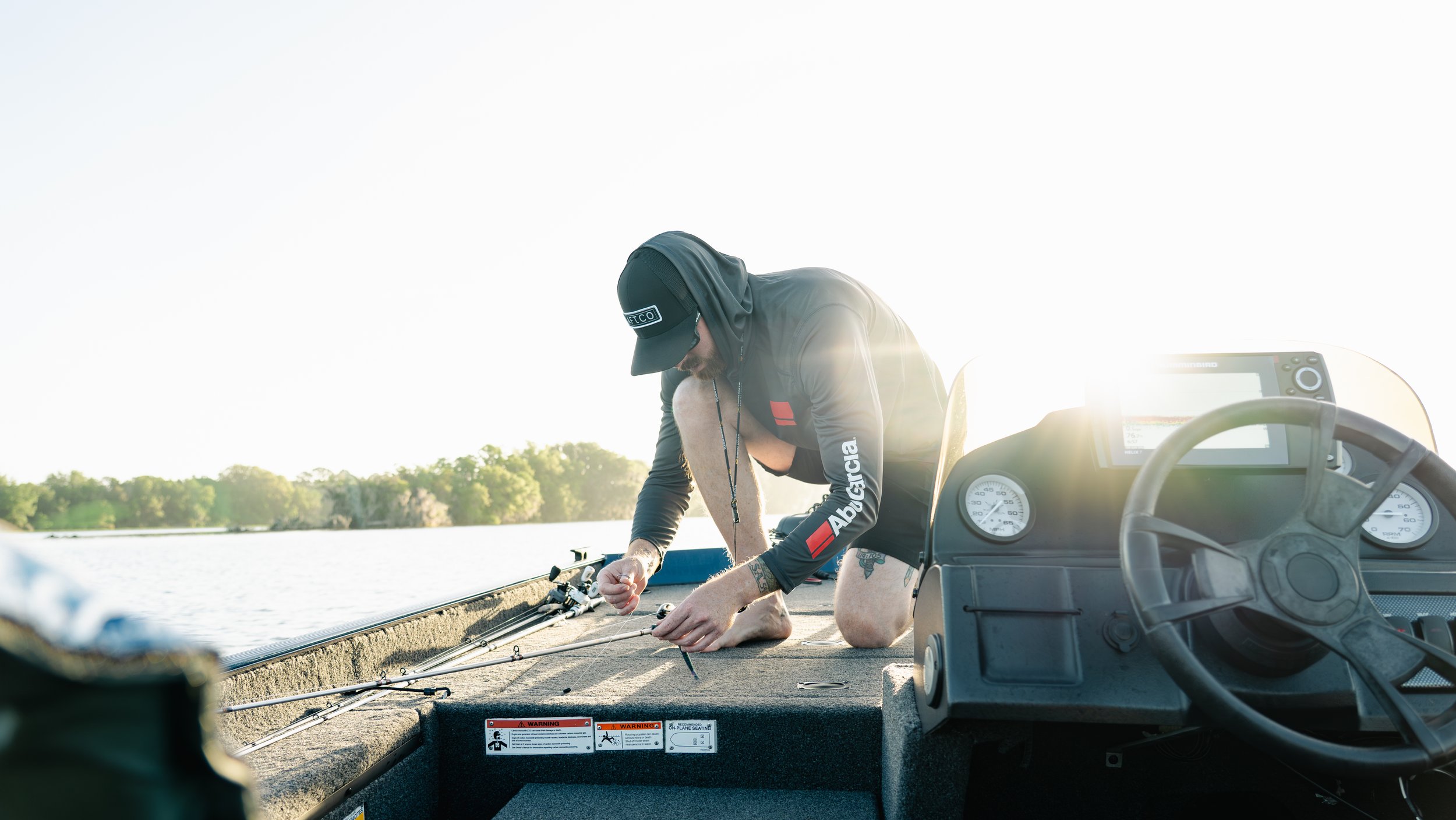Person kneeling on a boat, fishing rod in hand, wearing a black hoodie and cap, with boat dashboard and water in the background.