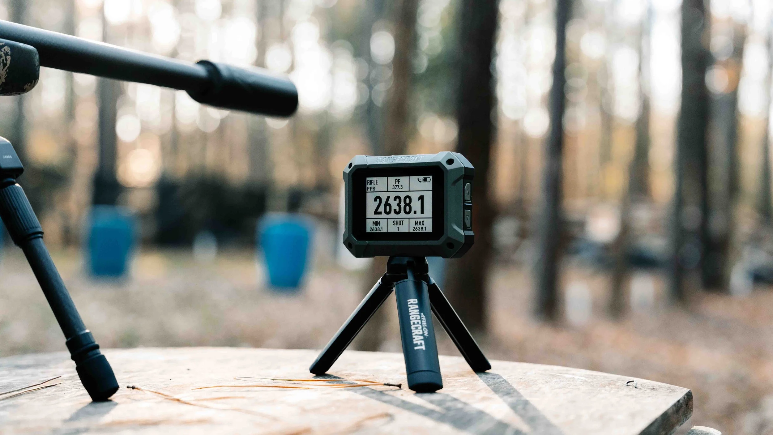 A rangefinder measuring distance on a wooden table outdoors in a forest with blue targets in the background.