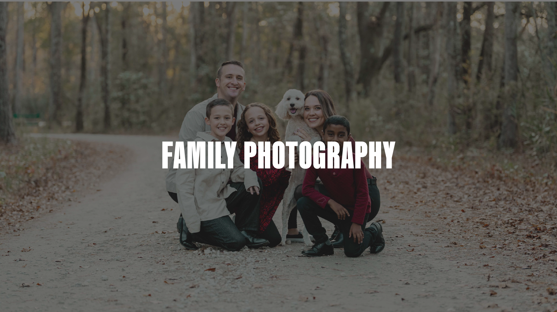 A family of six, including an adult man and woman, three children, and a white dog, gathered on a dirt path in a wooded area during fall, smiling at the camera with trees and fallen leaves surrounding them.