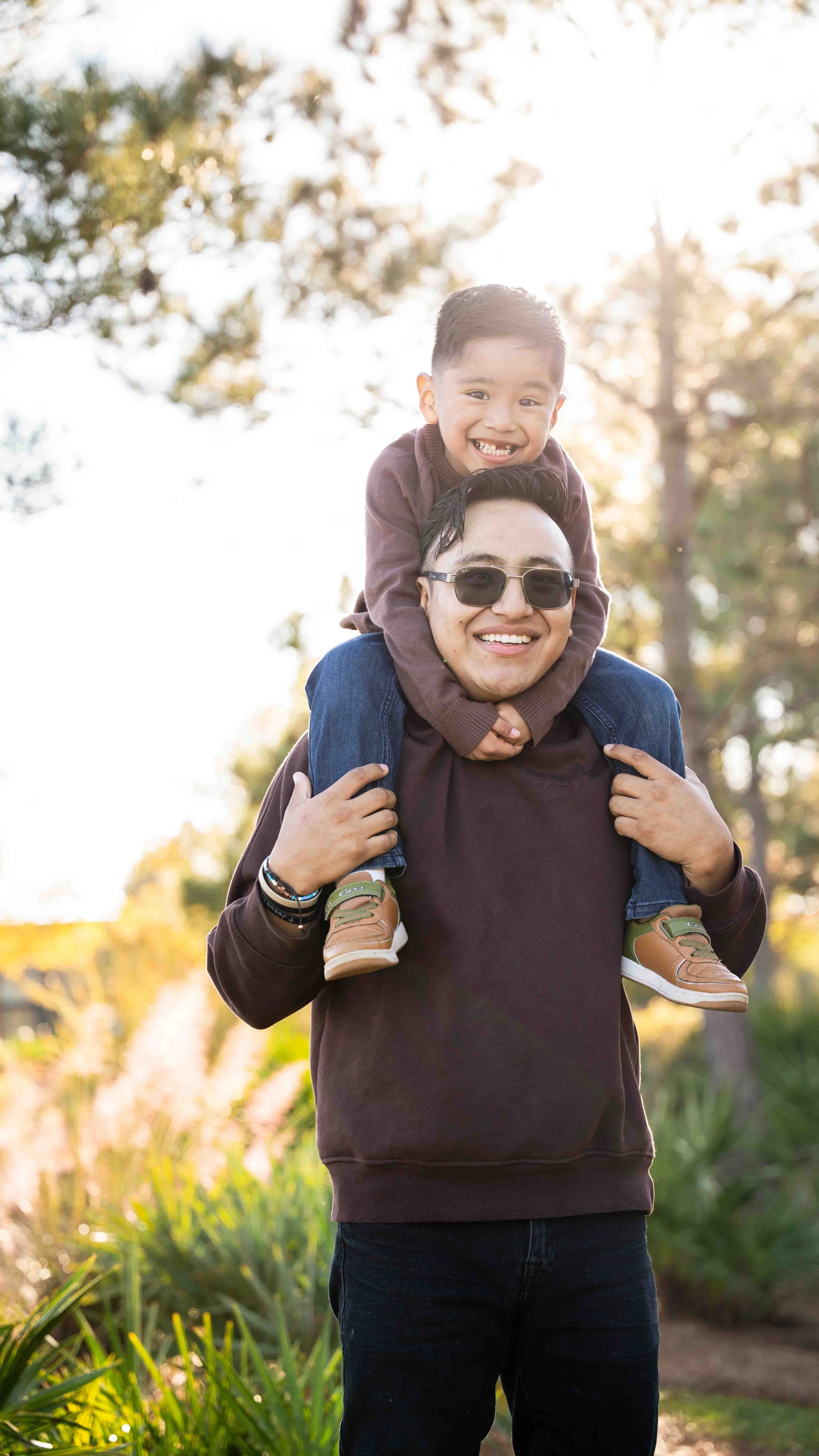 A man wearing sunglasses carrying a boy on his shoulders outdoors in a park or garden during sunset, with trees and greenery in the background.