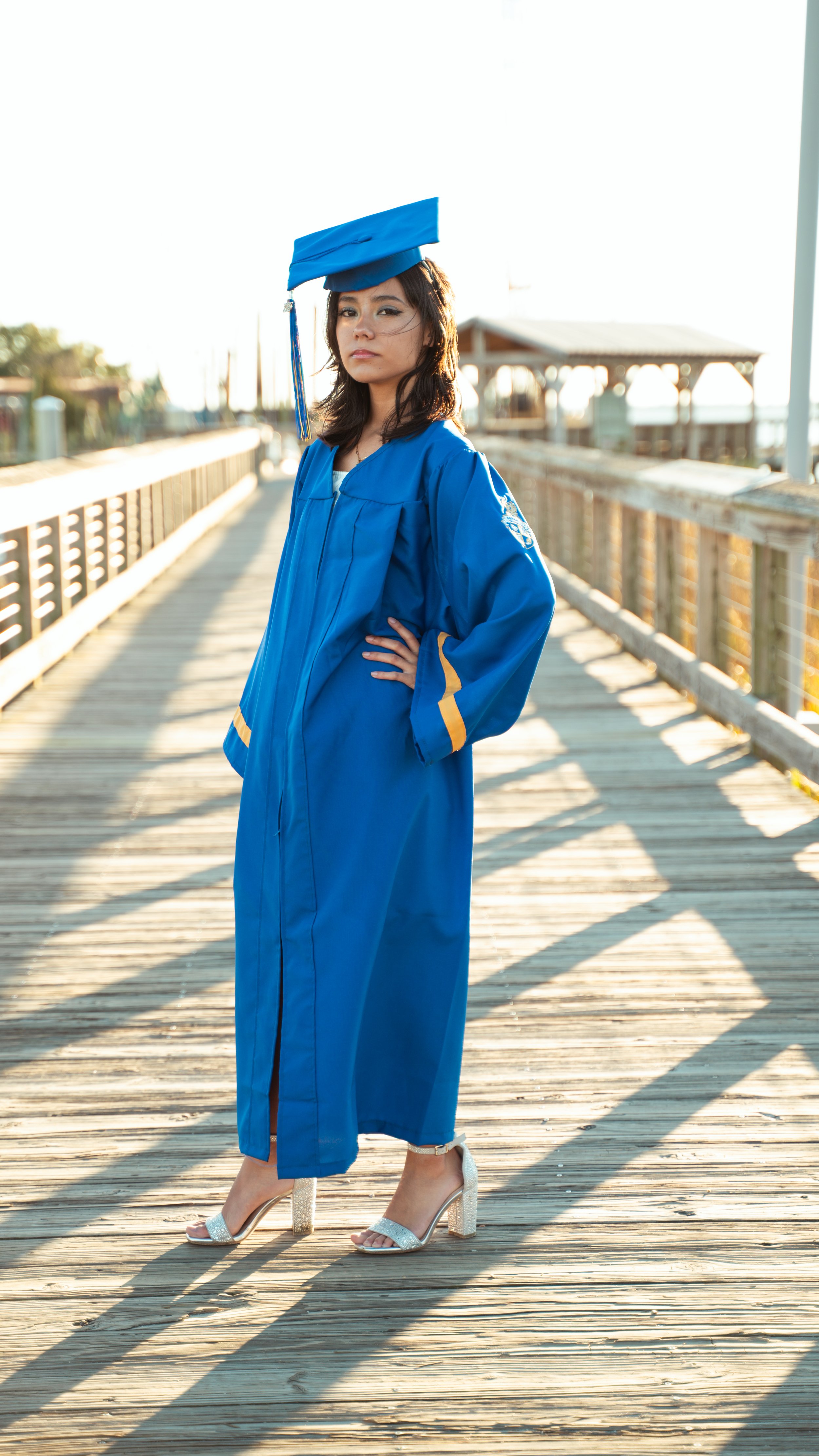 Young woman in a blue graduation gown and cap standing on a wooden pier during sunset, with her hands on her hips and looking at the camera.