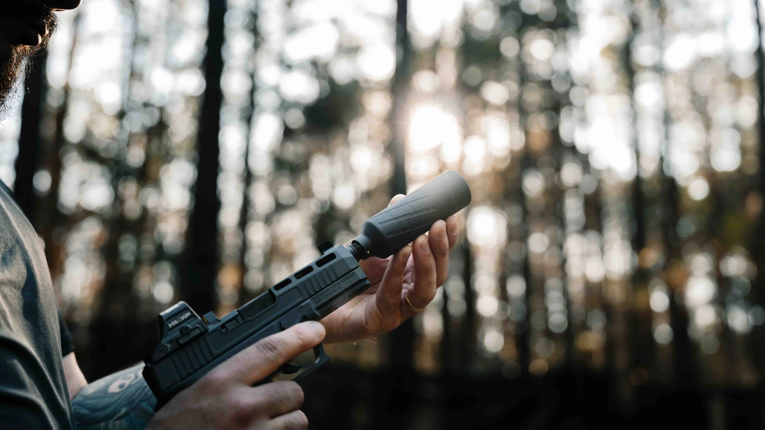 A person holding a handgun with a suppressor attached, outdoors in a wooded area with sunlight filtering through trees in the background.