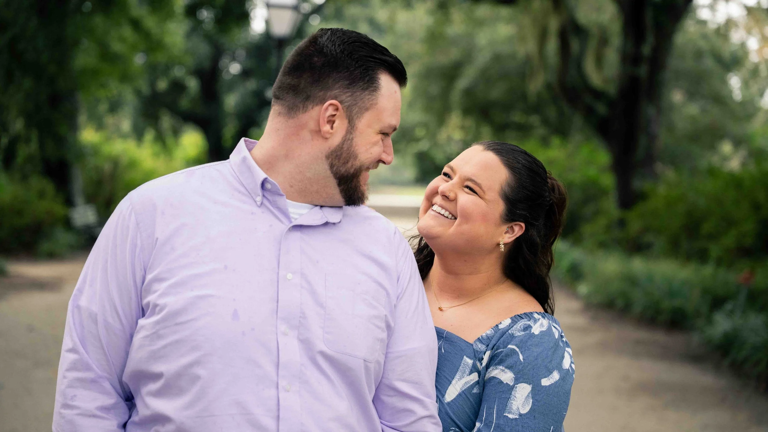 A couple smiling at each other outdoors in a park with trees and greenery in the background.