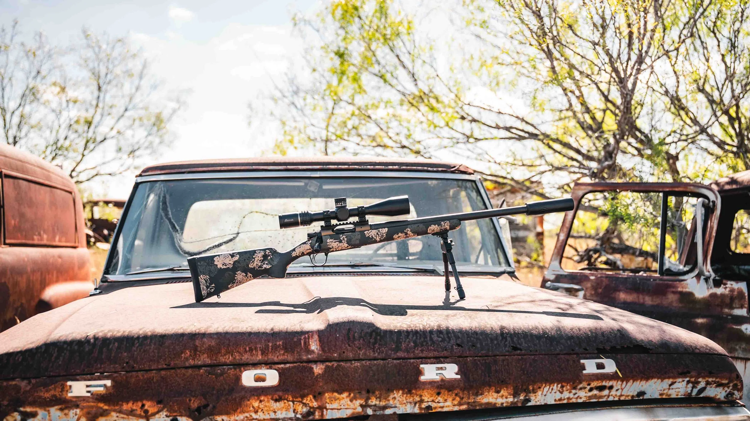 A rifle with a floral pattern on its stock and a scope mounted on top, resting on the hood of a rusted, old Ford vehicle in an outdoor setting with trees in the background.