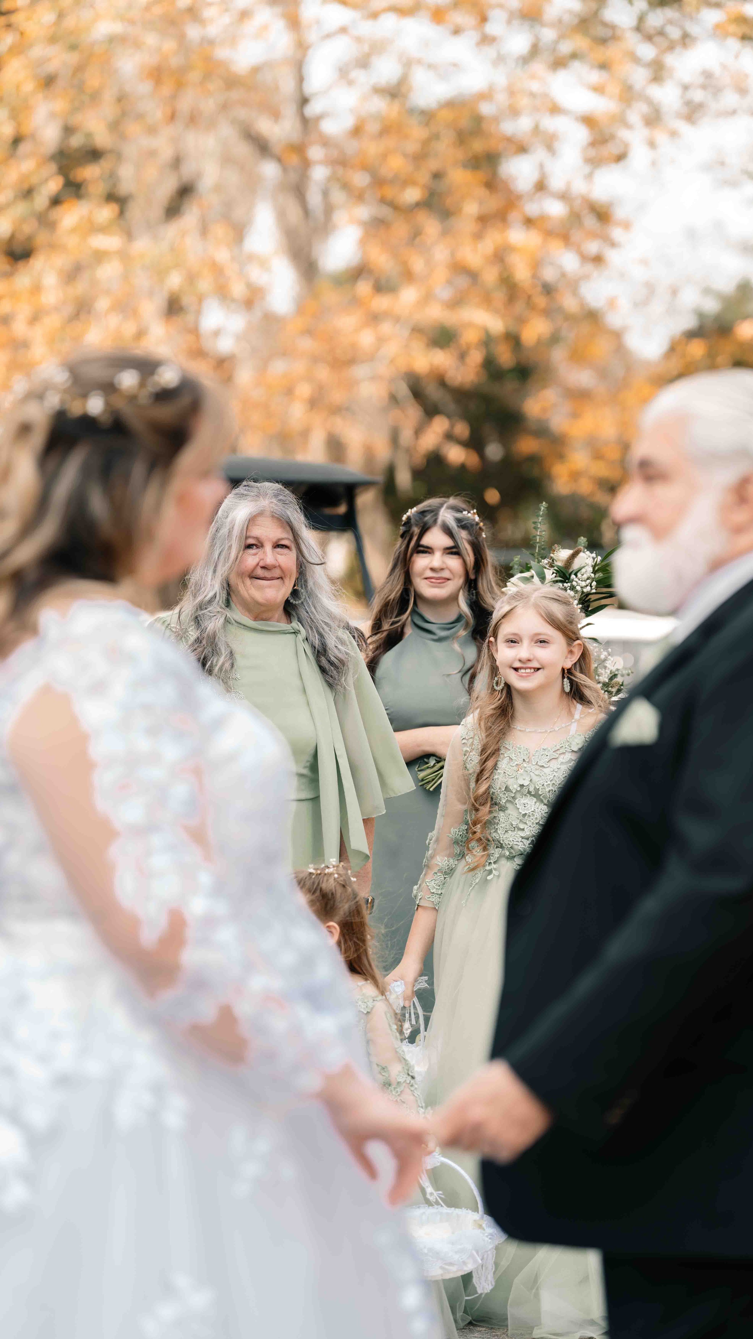 A wedding ceremony outdoors with the bride and groom holding hands, surrounded by women and a young girl, with autumn leaves on the trees in the background.