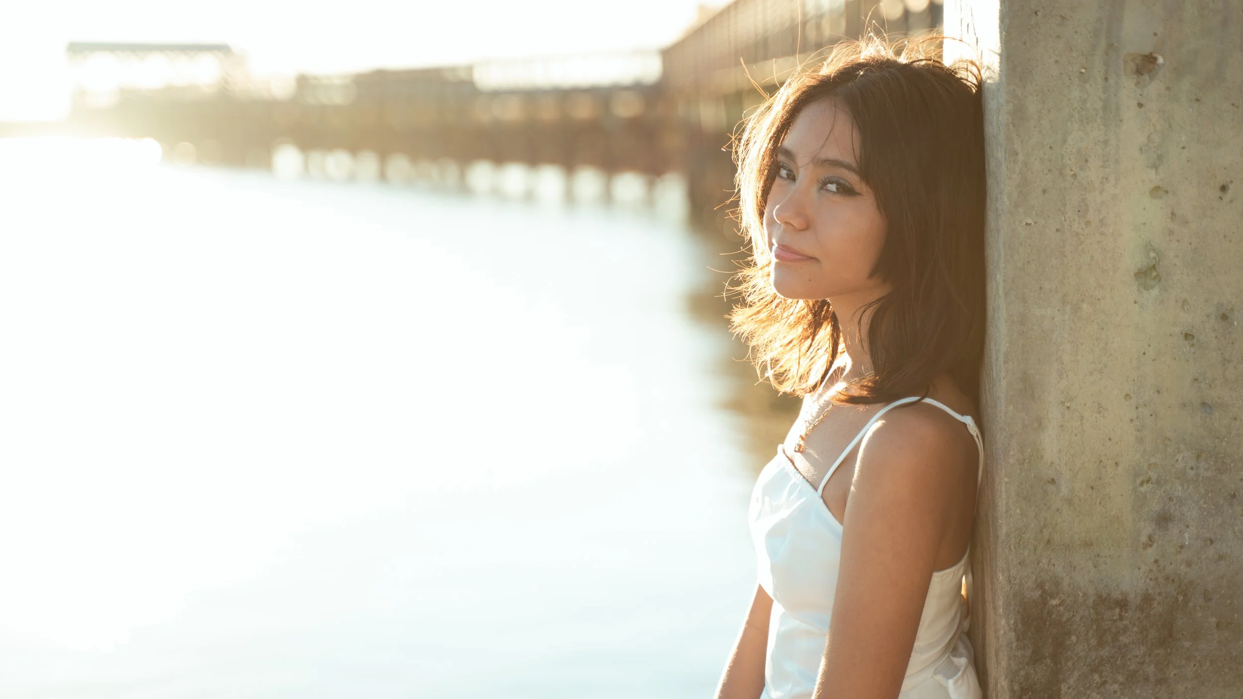 A woman leaning against a concrete pillar by a river during sunset, wearing a white spaghetti strap top, with shoulder-length brown hair, looking at the camera with a slight smile.