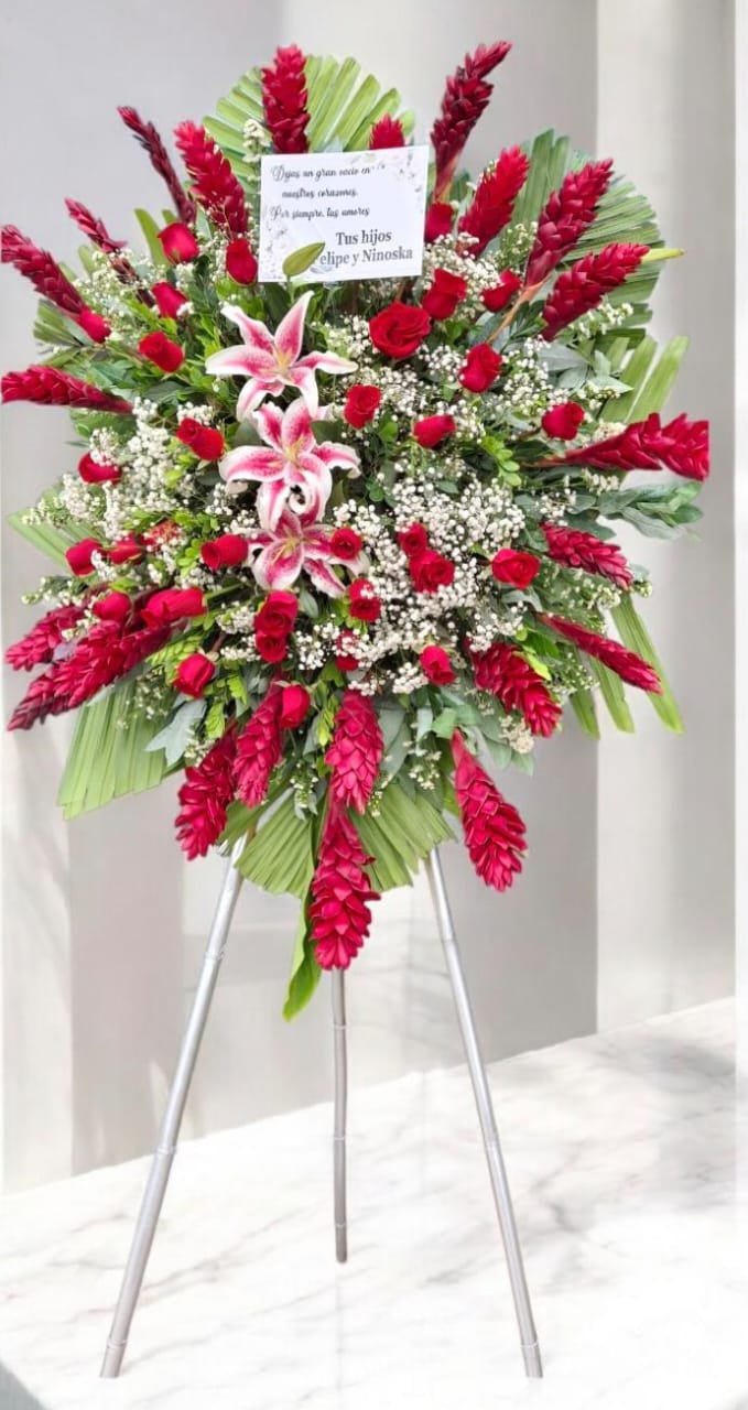 Arreglo floral con rosas rojas, lirios rosados, y flores blancas con follaje verde, en un soporte con tres patas.