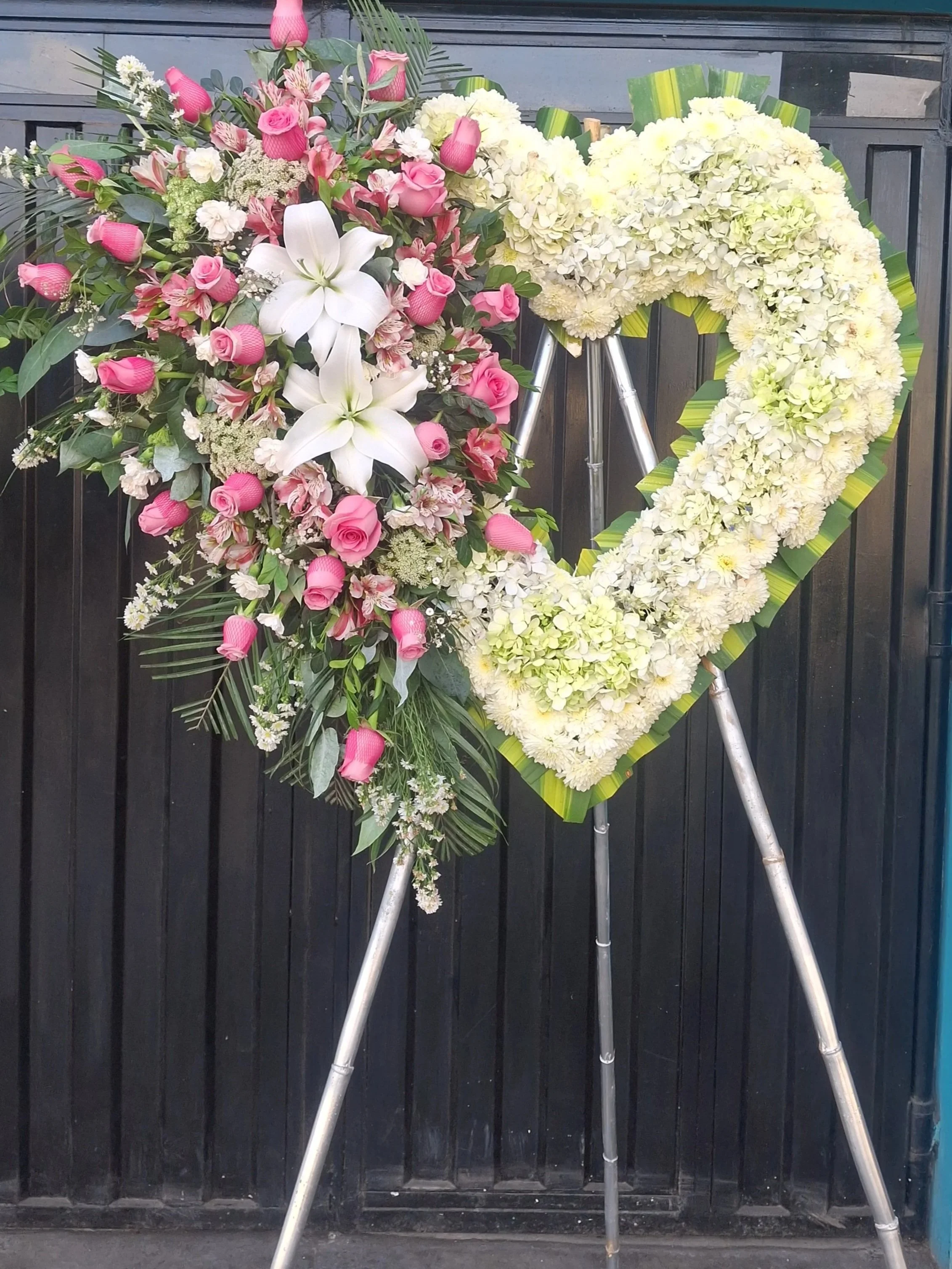 Arreglo floral con flores blancas y rosas, en forma de corazón, colocado sobre un trípode de metal, frente a una cerca de madera negra.