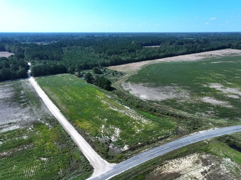 Aerial view of farmland with green fields, dirt roads, and a forest in the background under a blue sky with clouds. Open lots for modular, mobile, stick-built, or starter homes.