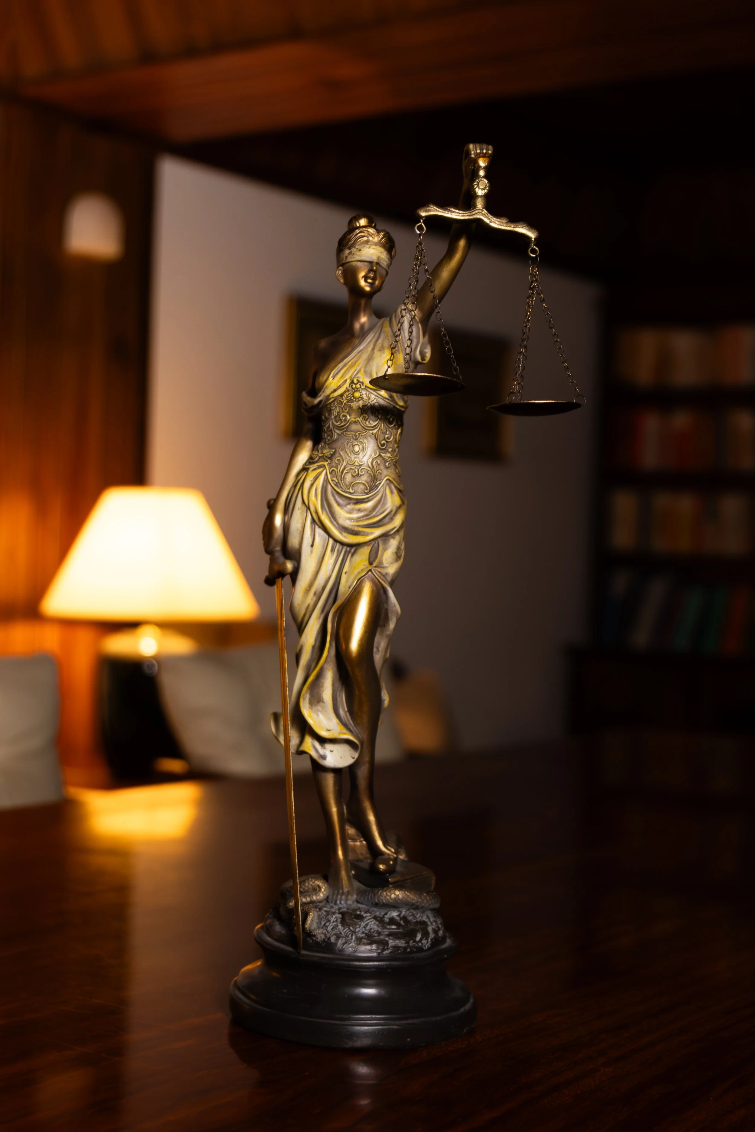 Estátua de uma mulher segurando uma balança, de pé sobre uma base decorativa, em um ambiente de iluminação baixa com uma luminária acesa ao lado. Fernando Anastácio e Associados