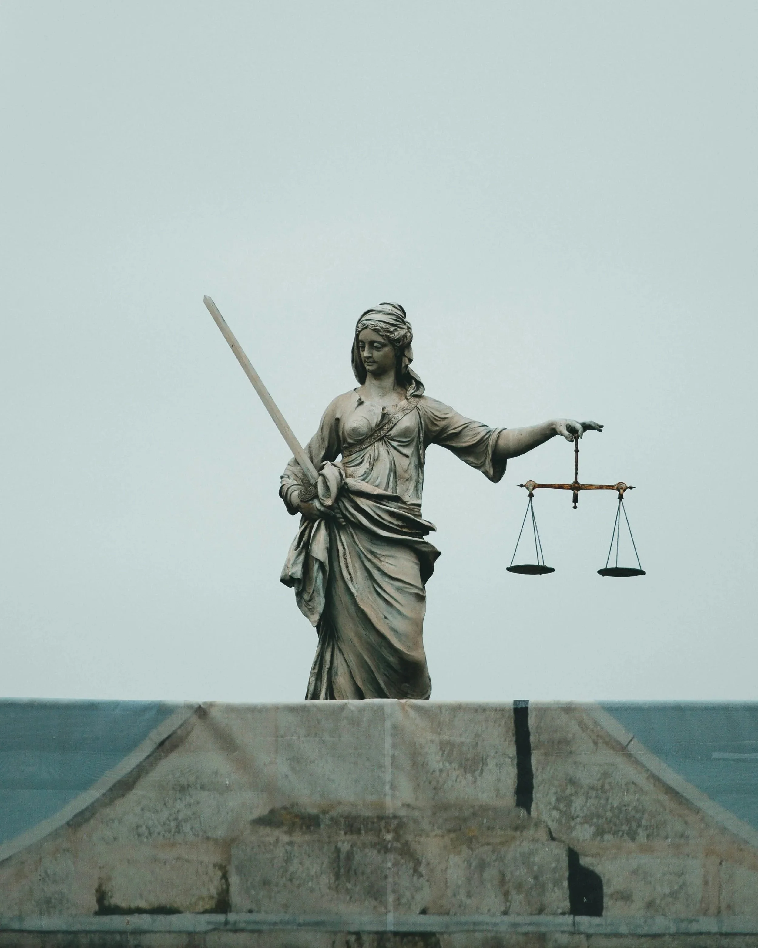 Estátua da deusa Tália segurando balança, com uma lança apoiada na mão esquerda. A estátua é de cor dourada e está em um pedestal, visto de baixo para cima contra um céu nublado. Fernando Anastácio e Associados