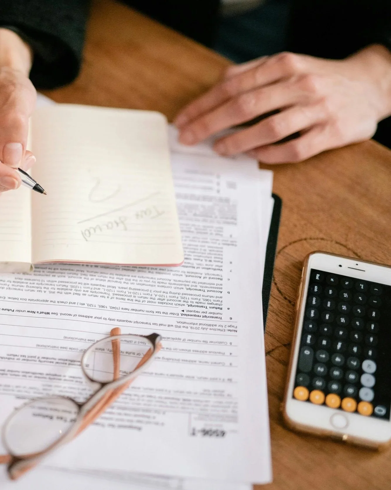 Mesa de escritório com documentos, óculos de grau, telefone celular com teclado numérico e anotação à mão em caderno. Fernando Anastácio e Associados