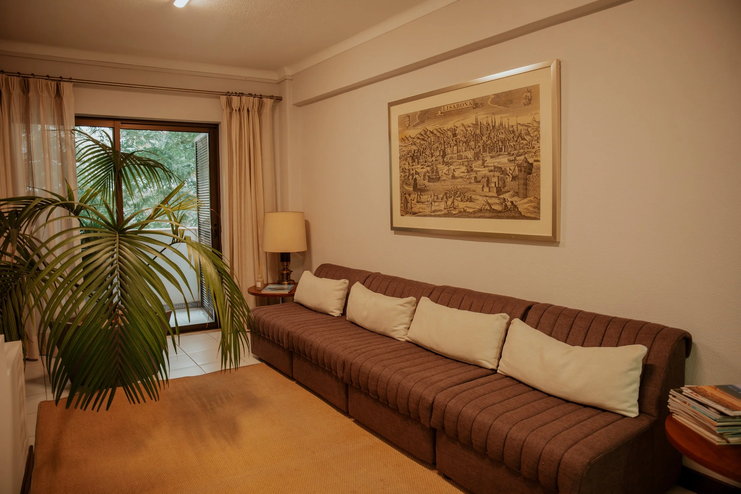 Sala de estar com sofá marrom com almofadas bege, janela de vidro com cortinas bege, planta de folhas longas, mesa de canto com revistas, quadro decorativo na parede, luminária ao lado do sofá, tapete bege. Fernando Anastácio e Associados