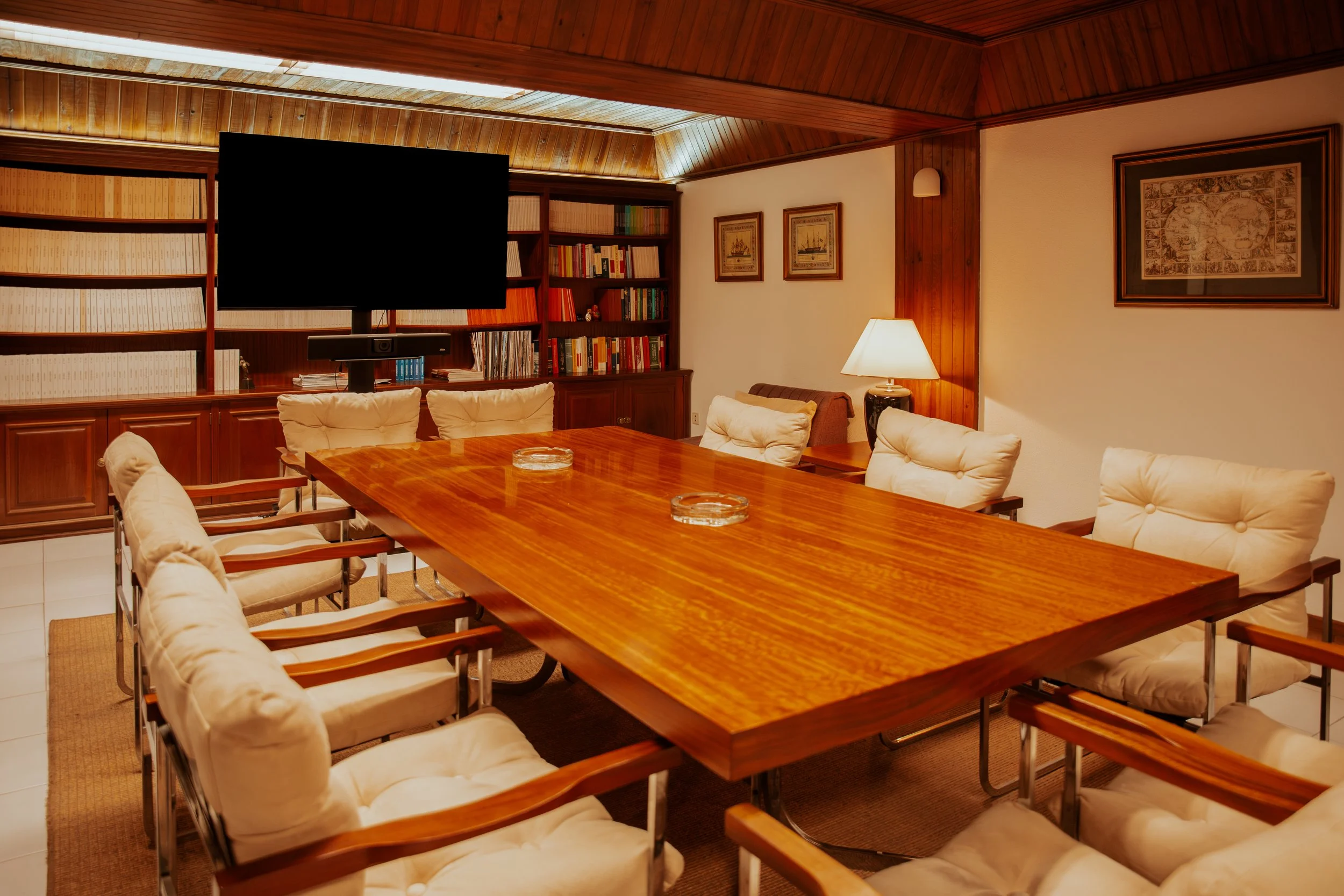 Sala de reunião com mesa de madeira e cadeiras com almofadas brancas, estante de livros, televisão e quadro na parede. Fernando Anastácio e Associados