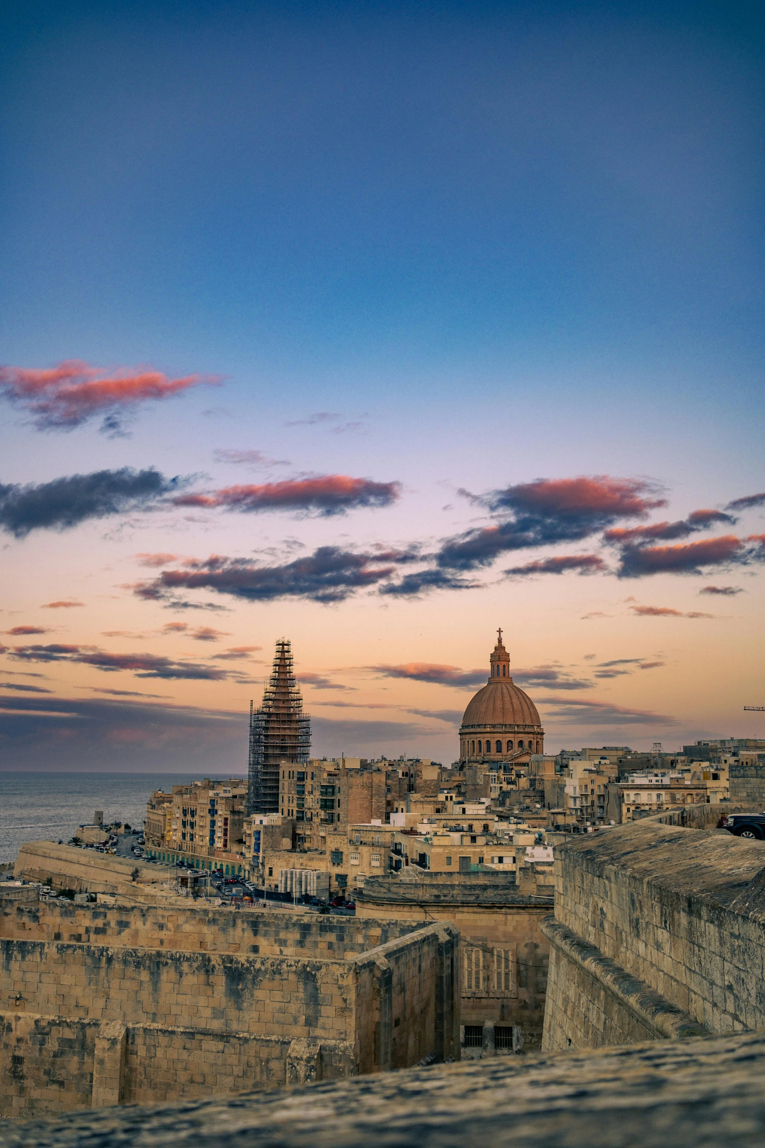Cena de uma cidade histórica ao pôr do sol, com cúpulas e edifícios antigos ao lado do mar.