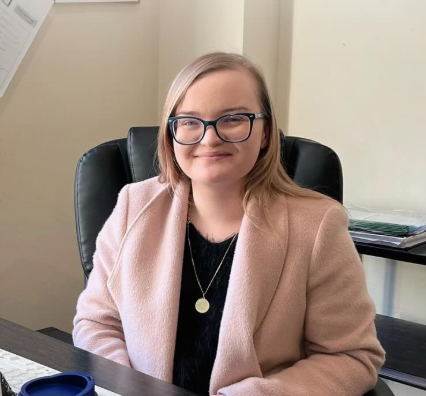Mulher sorrindo, usando óculos e blazer rosa claro, sentada em uma mesa de escritório com papelada ao fundo.