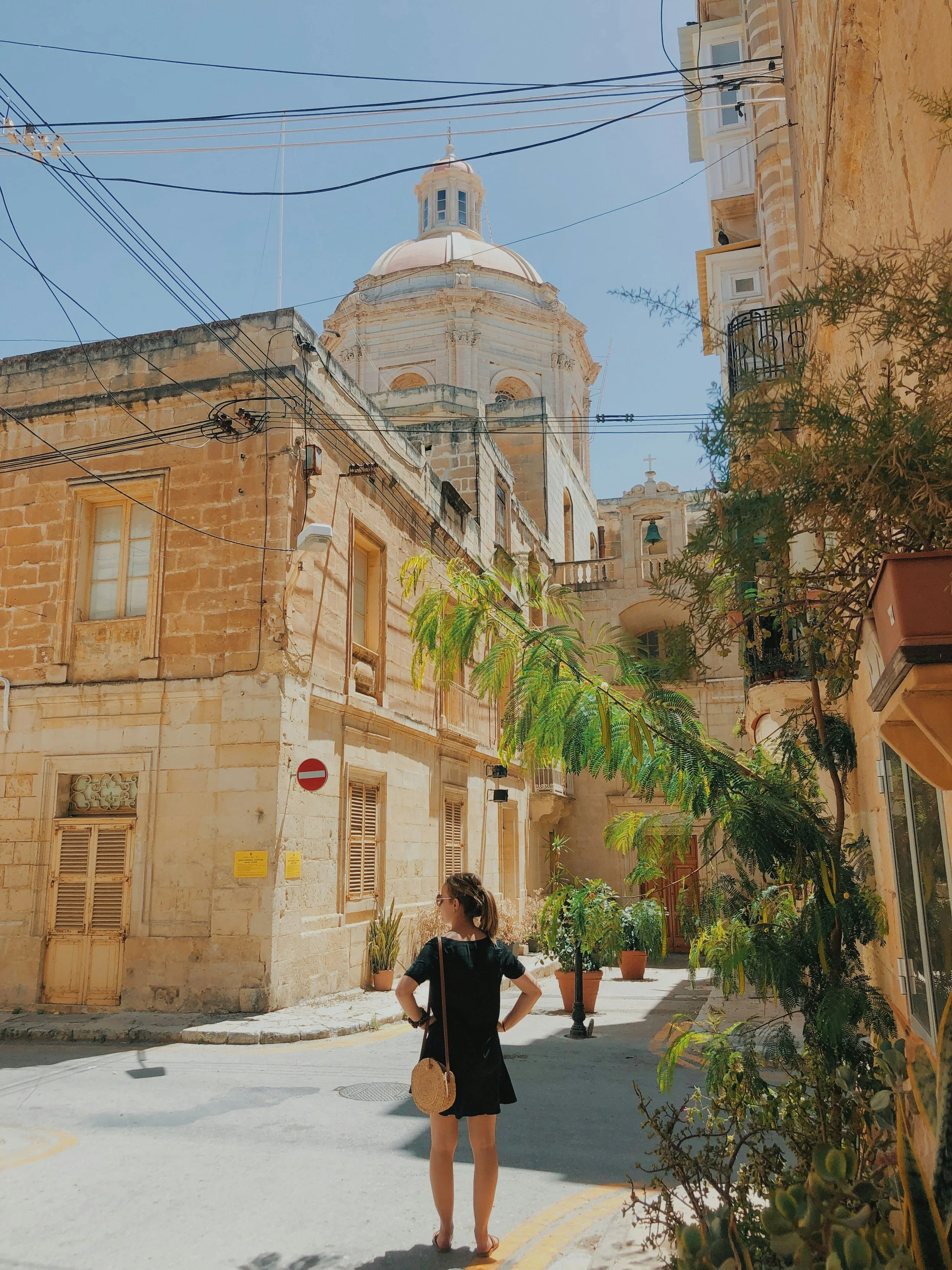 Mulher de vestido preto e bolsa de palha caminhando por rua estreita de cidade antiga, com edifícios de pedra e uma grande cúpula branca ao fundo, céu azul claro ao dia.