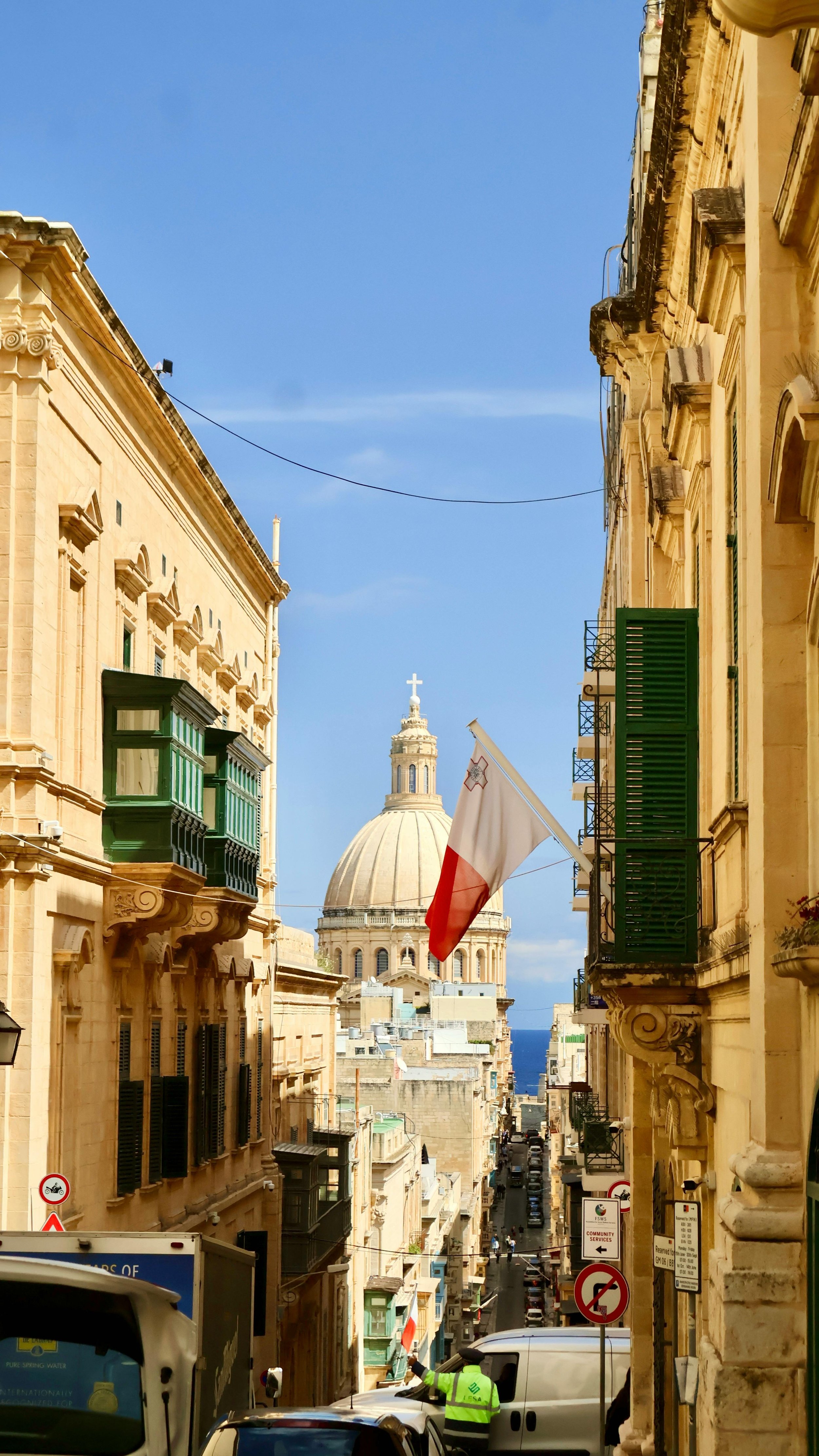 Rua estreita com edifícios antigos de ambos os lados, balões de ensaio presos às janelas, uma bandeira de Malta e uma do Vaticano no centro, com uma cúpula de igreja ao fundo e céu azul claro.