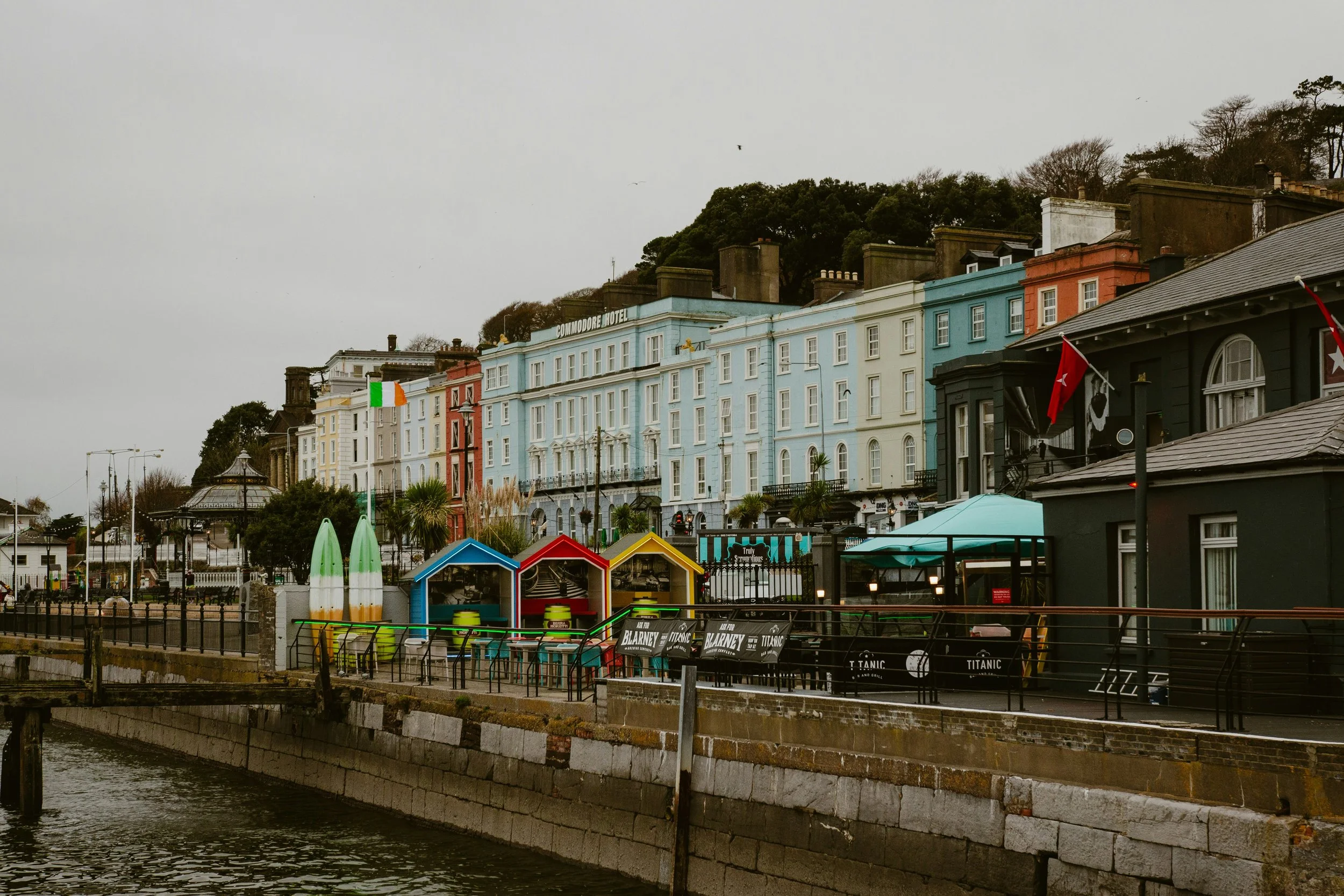 Casas coloridas ao longo de um cais com bancos e sinalizações, ao lado de um rio, com árvores ao fundo e céu nublado.