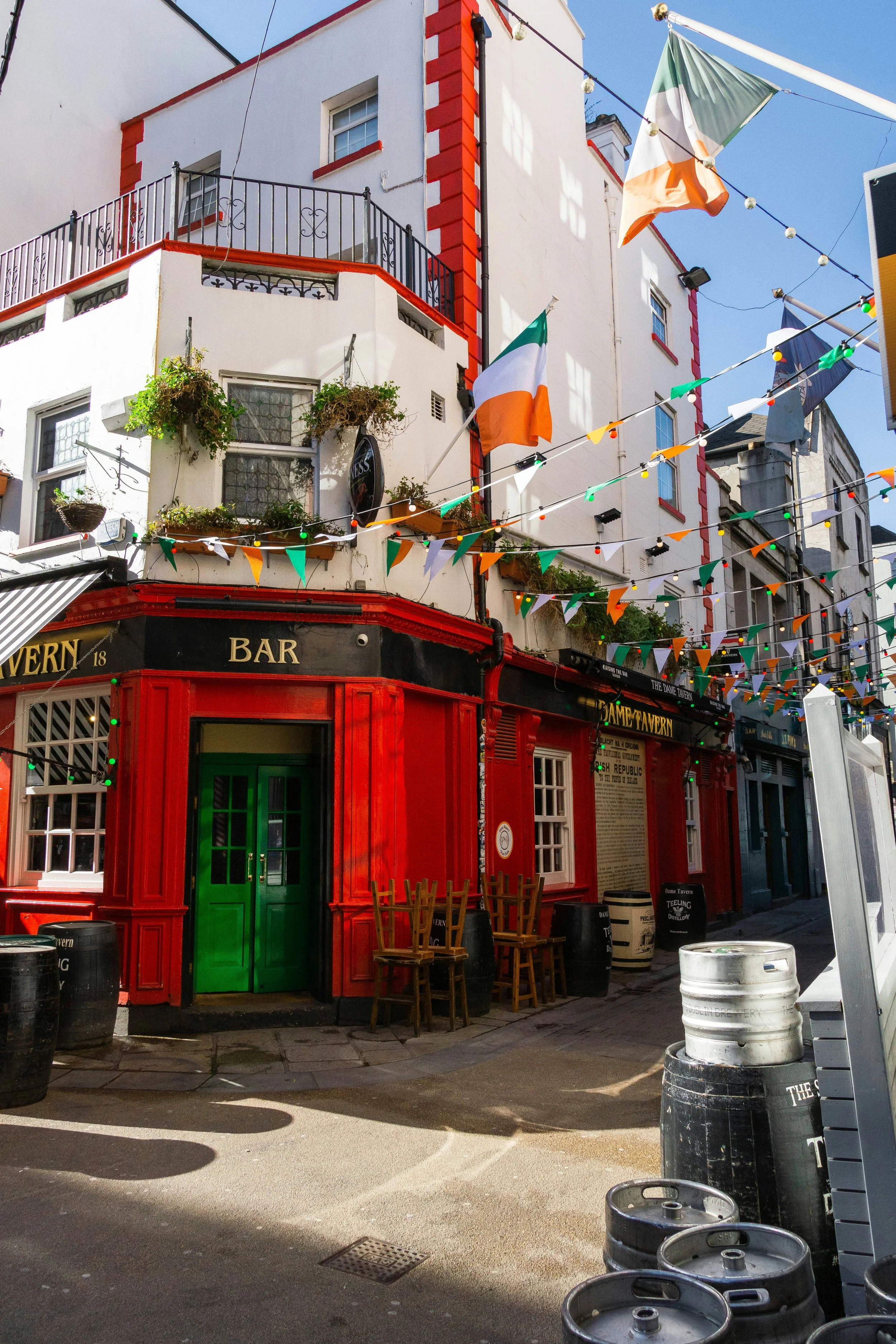 Bar com fachada vermelha, porta verde, cadeiras de madeira na calçada, bandeiras da Irlanda e decoração colorida no centro de uma rua