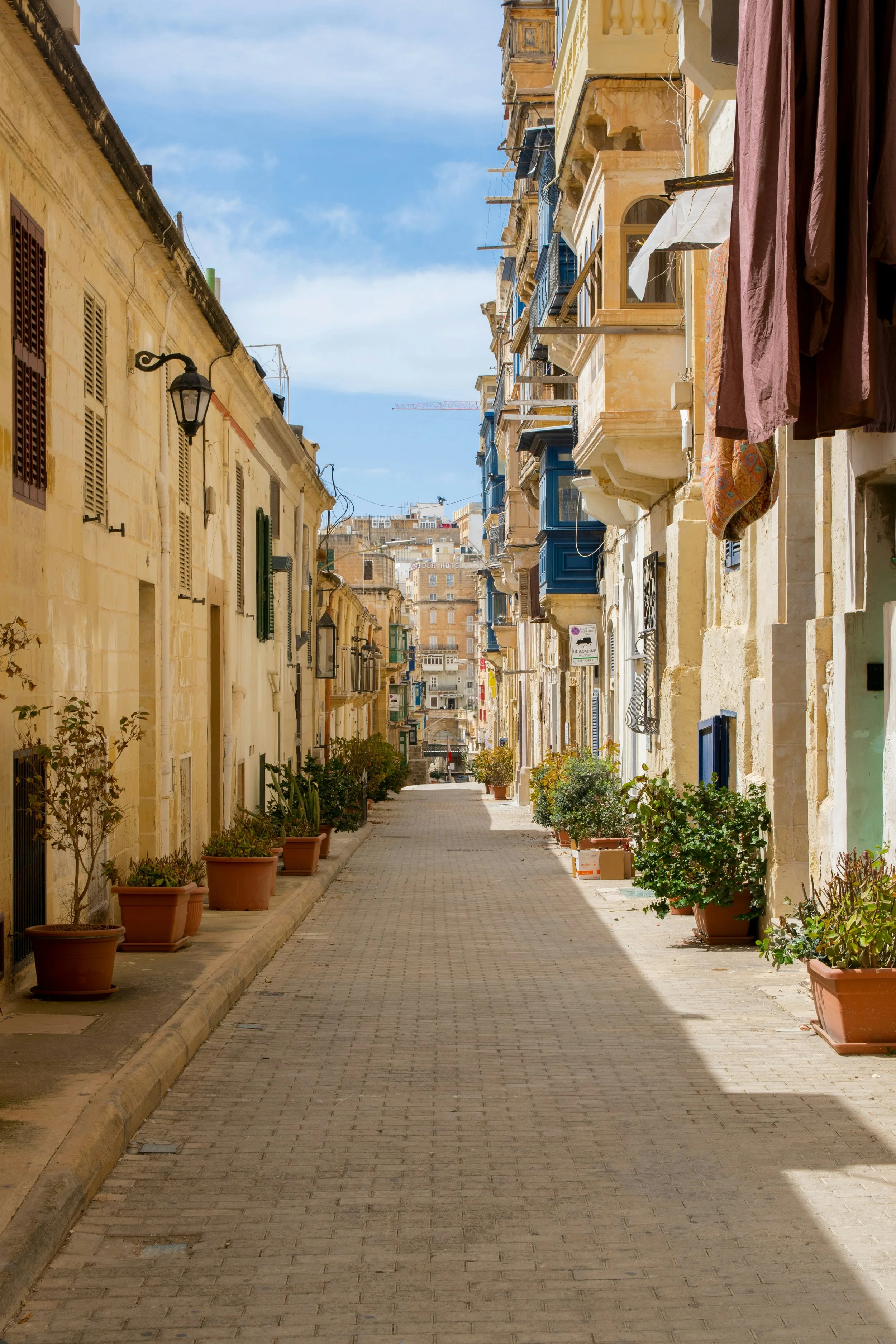 Rua estreita com calçada de pedras, ladeada por edifícios de fachadas de pedra, algumas com varandas e roupas penduradas para secar, com plantas em vasos ao longo da calçada, sob um céu azul com algumas nuvens.