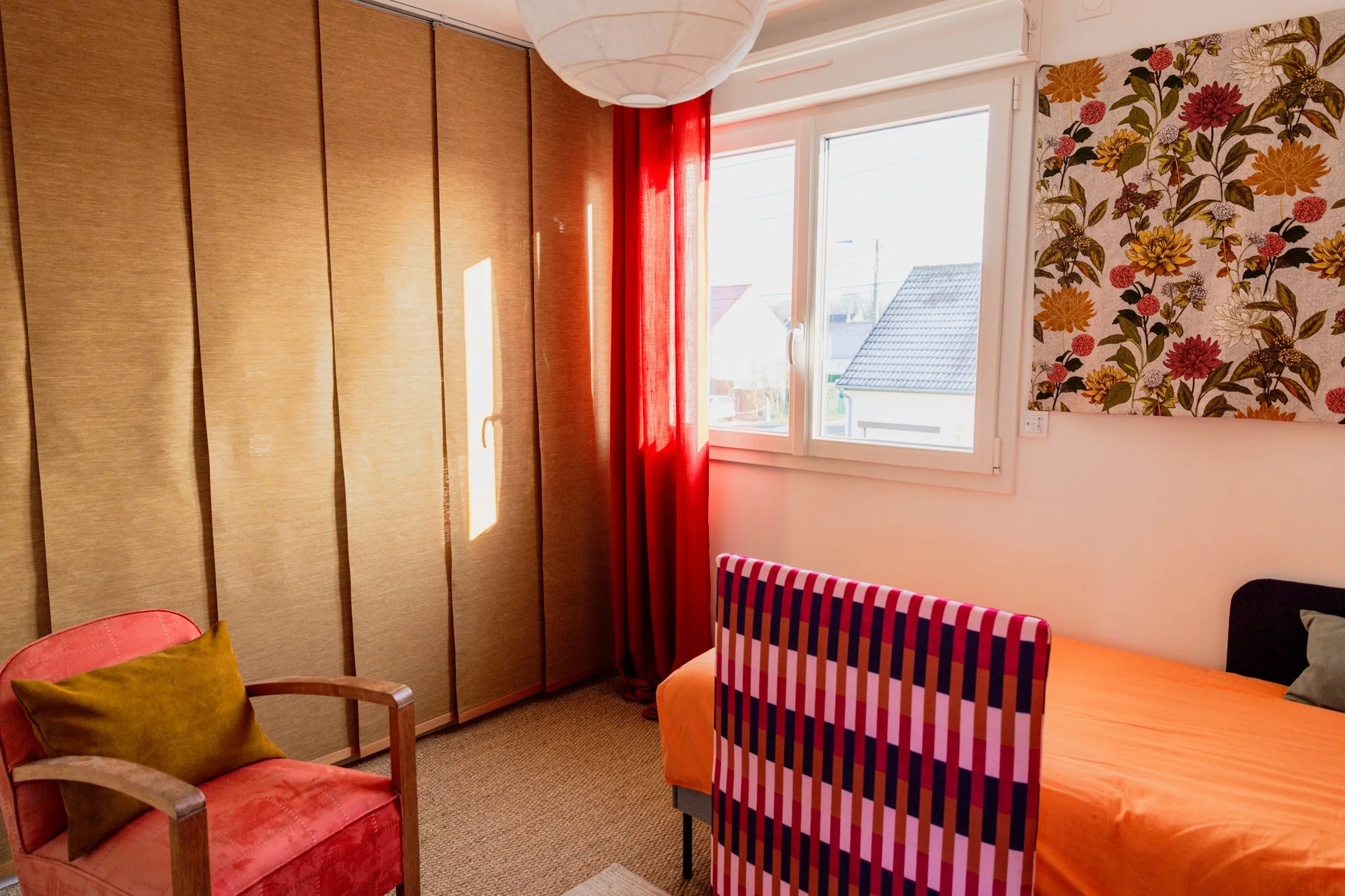 Colorful bedroom with orange bedding, striped chair, floral wallpaper, red curtains, and a sunlit window