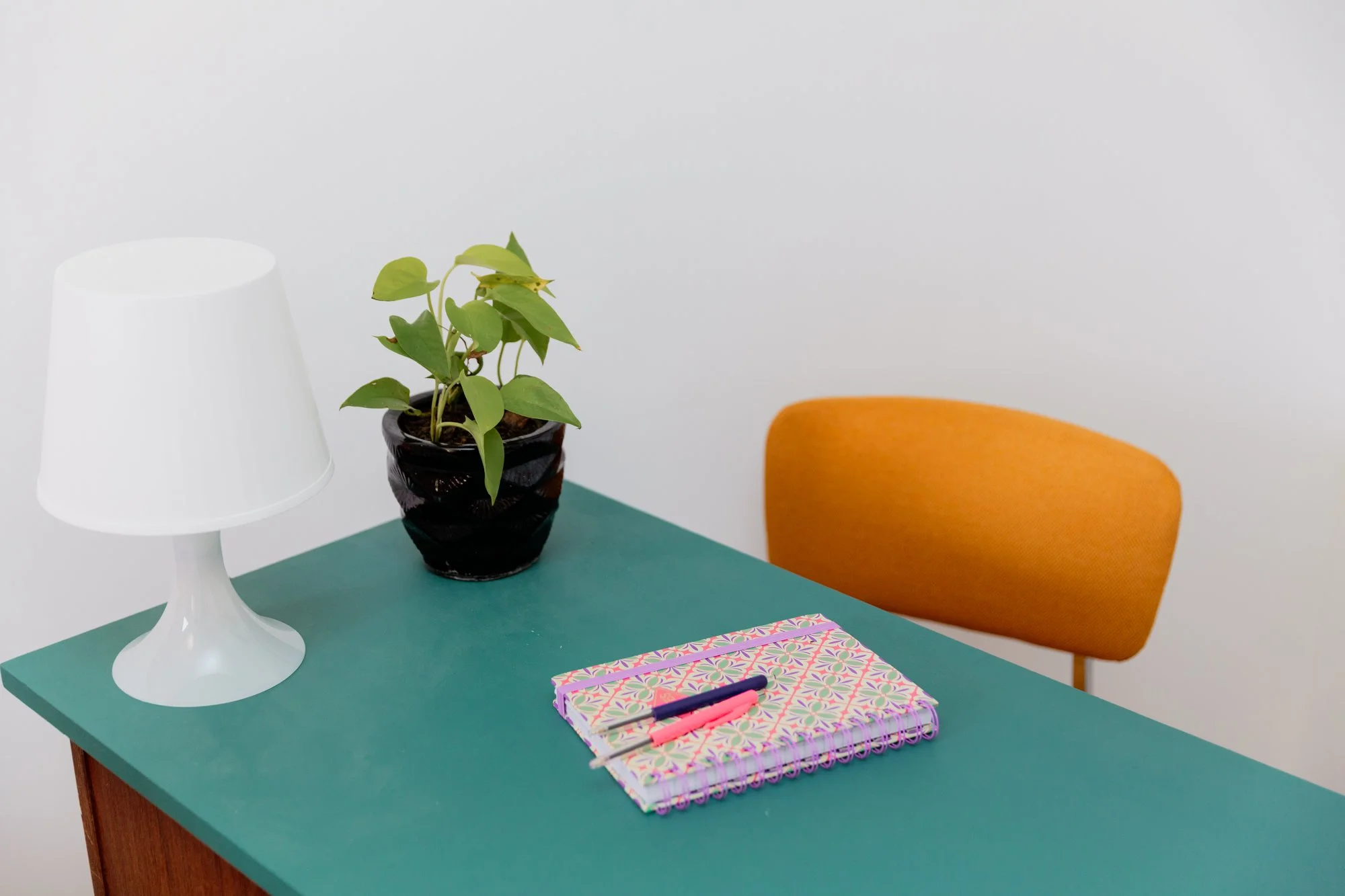 Colorful workspace with a white table lamp, potted green plant, patterned notebook, pens, and orange chair on a teal desk
