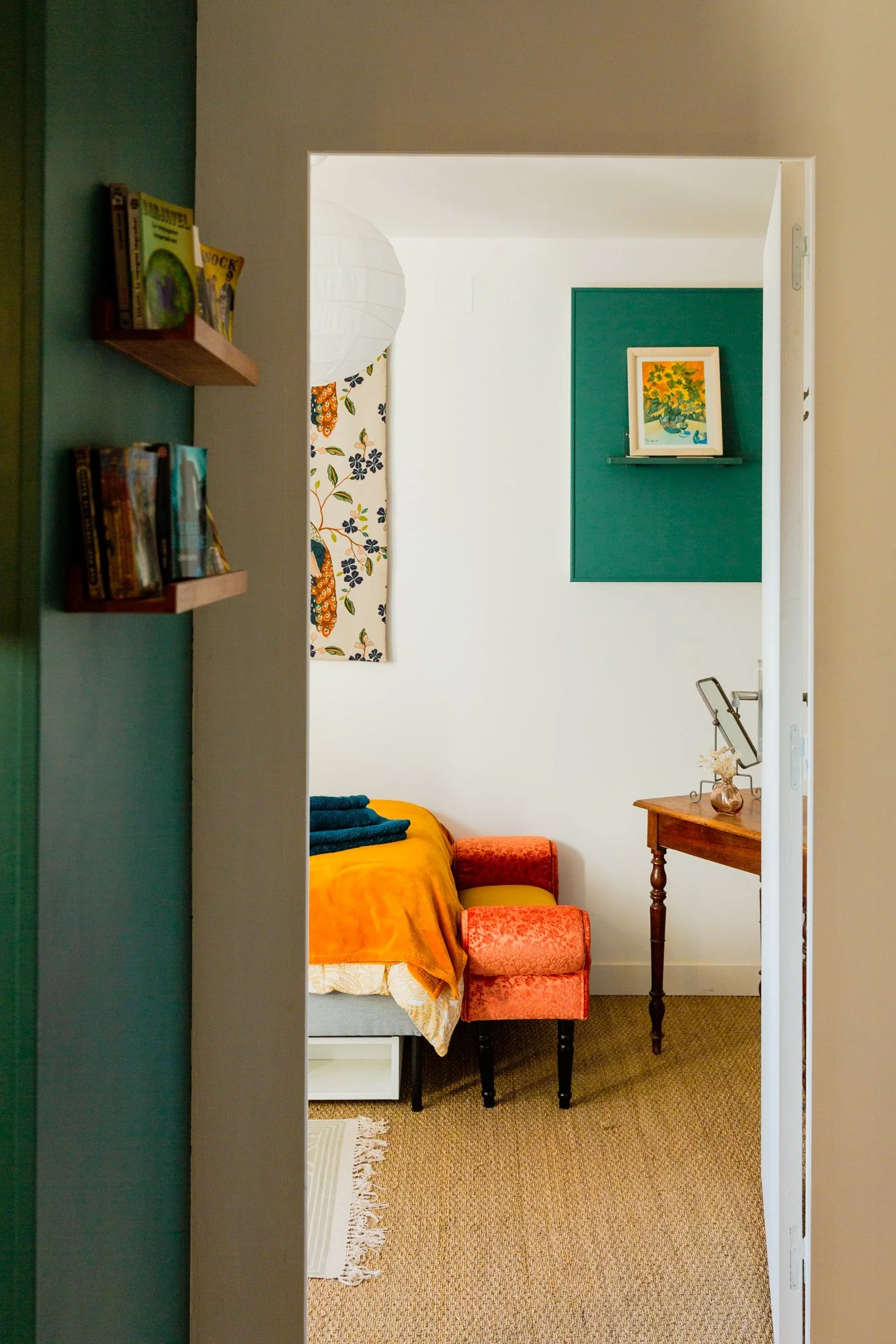 Colorful bedroom viewed through an open doorway, with bookshelves, orange bedding, patterned armchair, desk, and framed sunflower artwork.