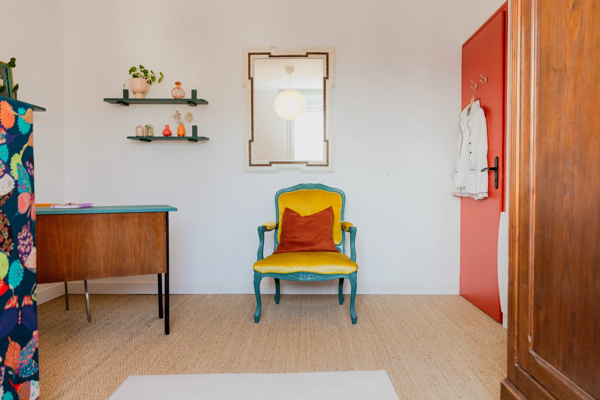 Colorful vintage-style room with a yellow upholstered armchair, red open door, wall mirror, floating shelves, and wooden cabinet.