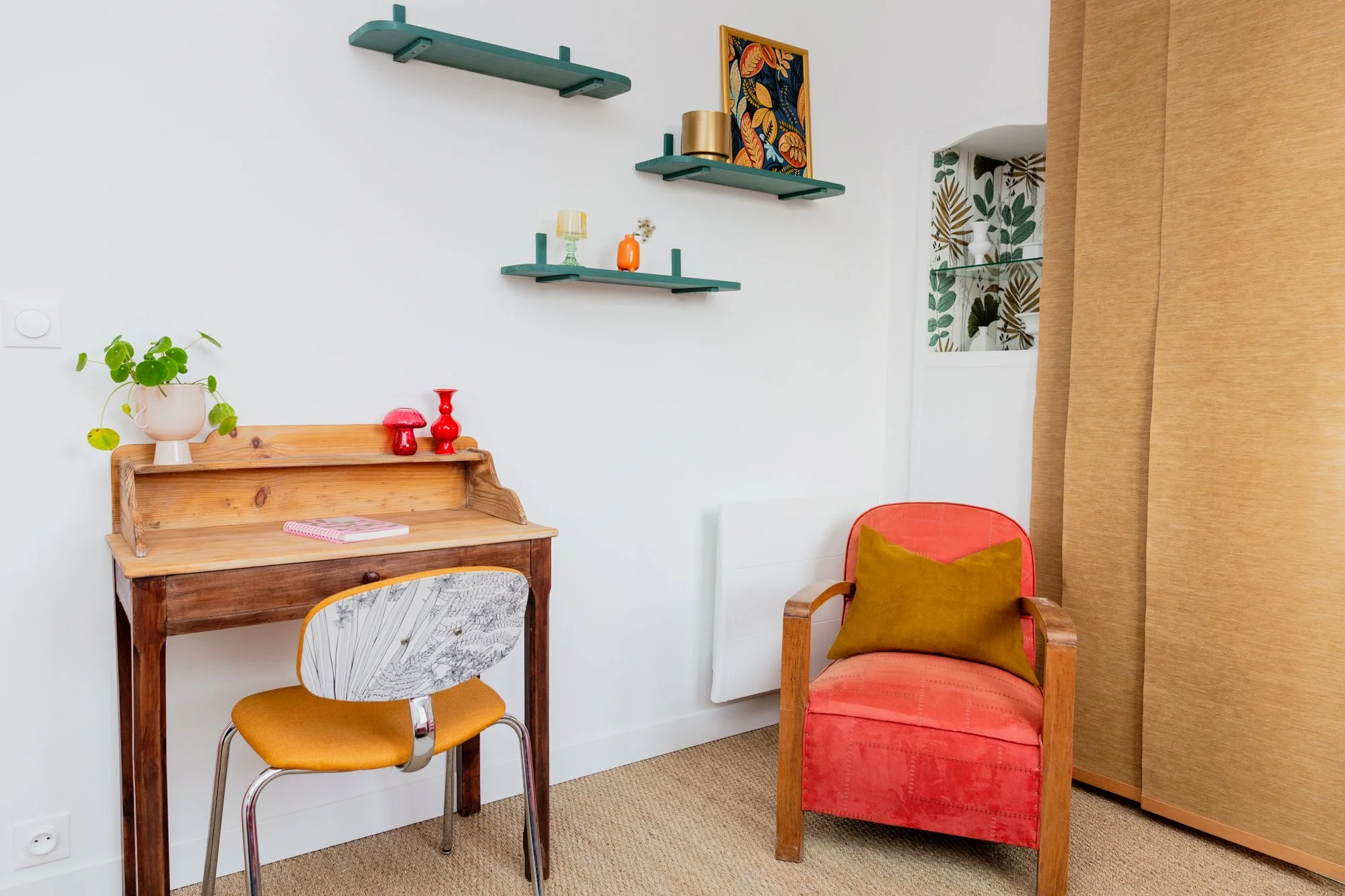 Colorful home office with a vintage wooden desk, mustard chair, coral armchair, wall shelves, and houseplants